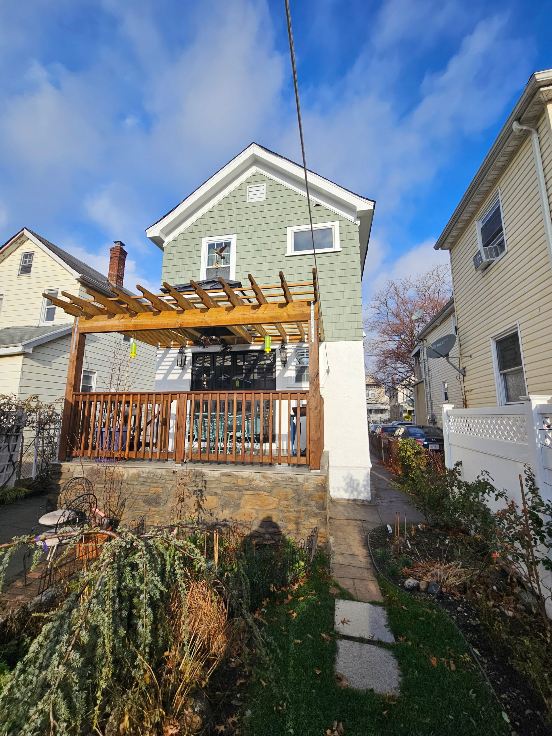 Two-story green house with a wooden deck and pergola, surrounded by a small yard with bushes and a walkway.