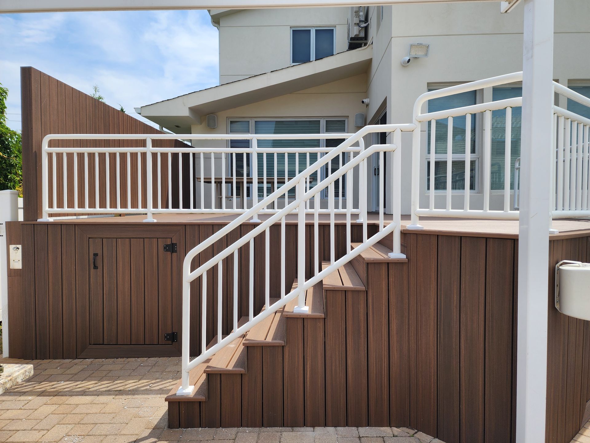 A white-railed deck with stairs leading up to a light-colored house with multiple windows. The deck is made of brown composite wood.