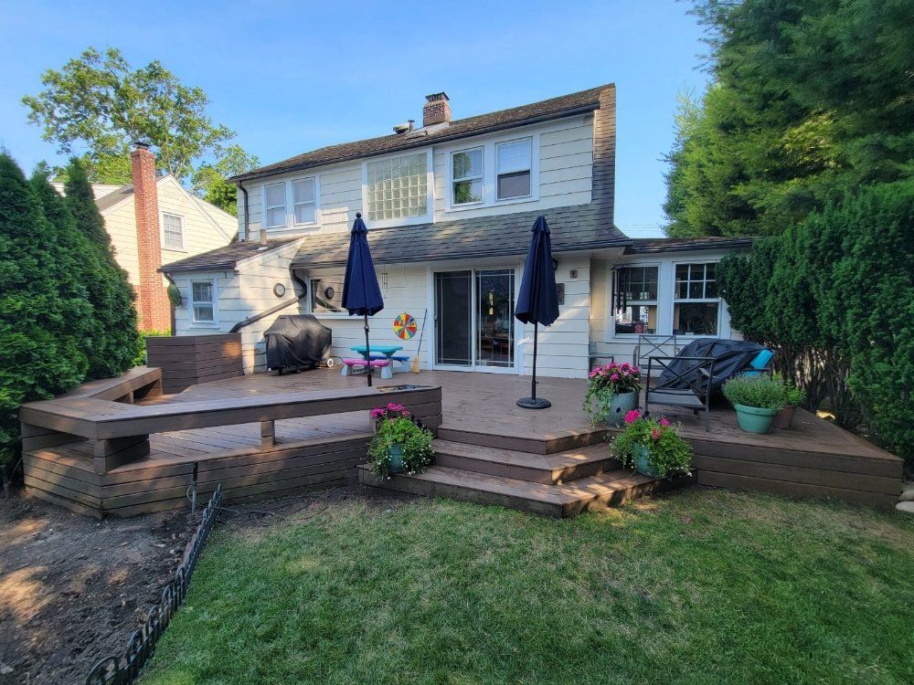 Backyard deck with a two-story house. The deck has stairs, built-in seating, and two blue umbrellas. Green grass and foliage surround the deck.