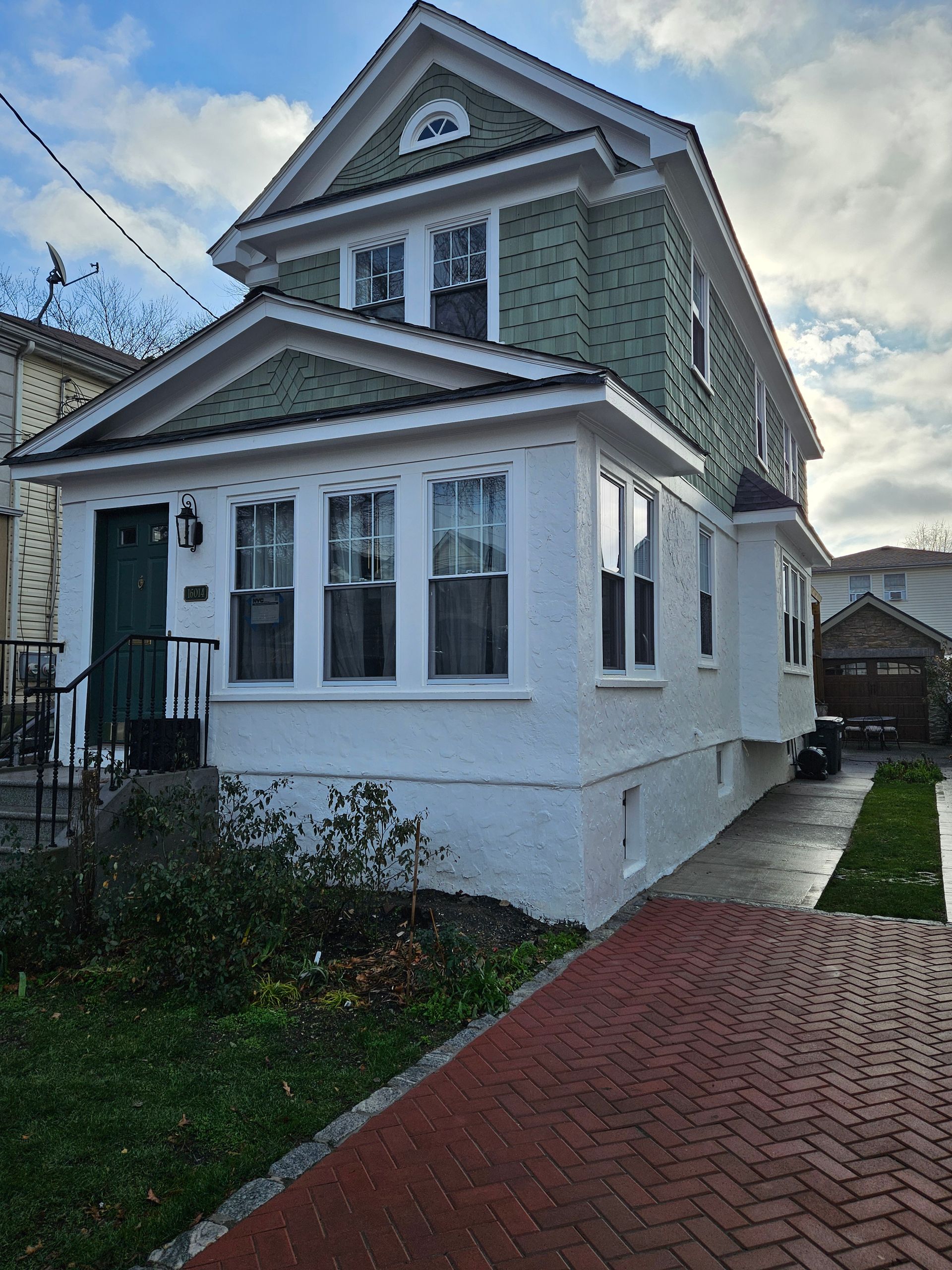Two-story house with a white stucco facade, green siding on the upper level, and a brick driveway. Cloudy sky in the background.