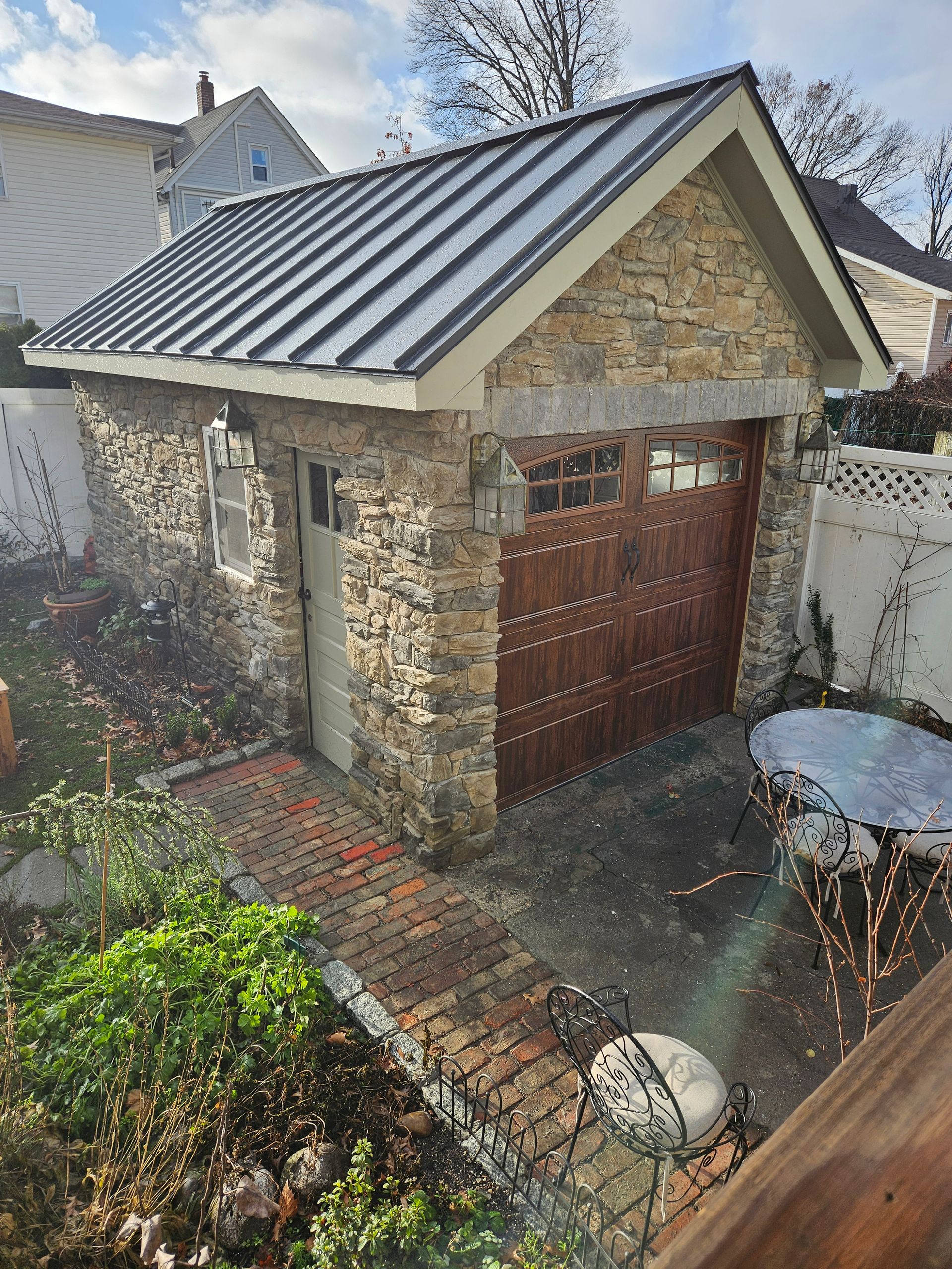 Stone garage with a brown wooden garage door and a metal roof. A brick path leads to the entrance.