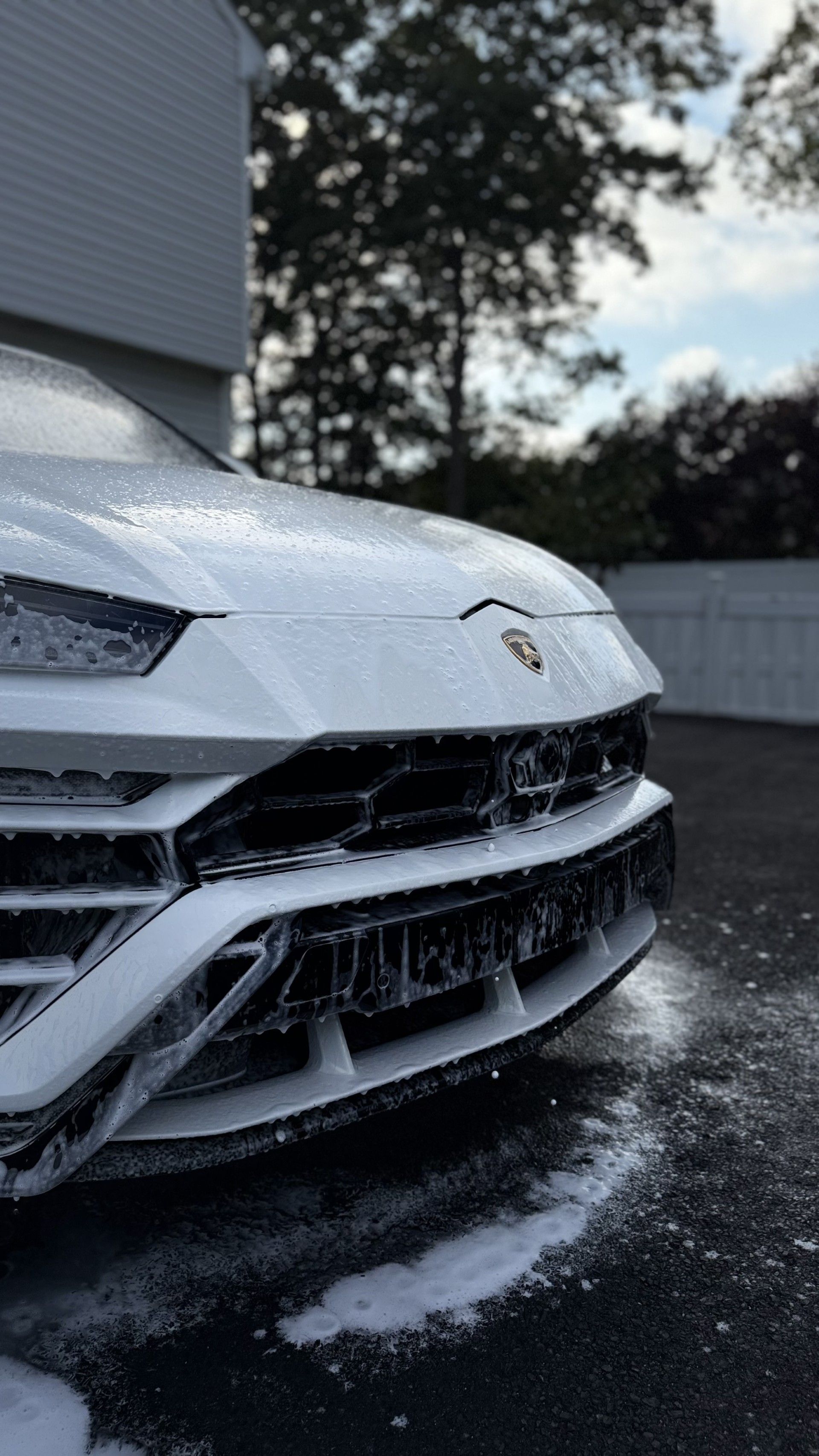 White sports car front bumper washing in a foamy spray outdoors near a building and fence