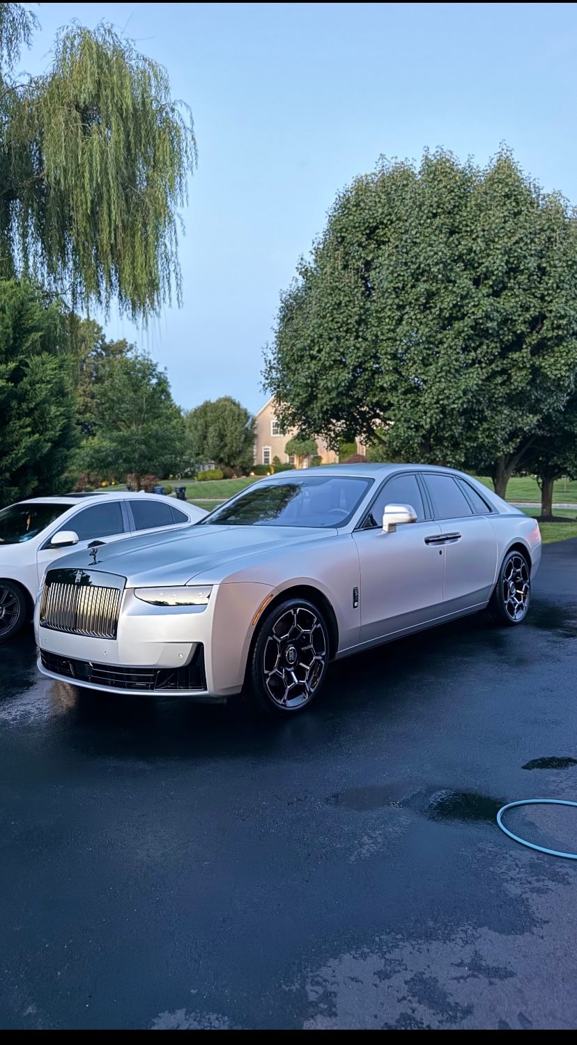 Silver Rolls-Royce parked on a wet driveway beside a white car, with trees and blue sky in the background