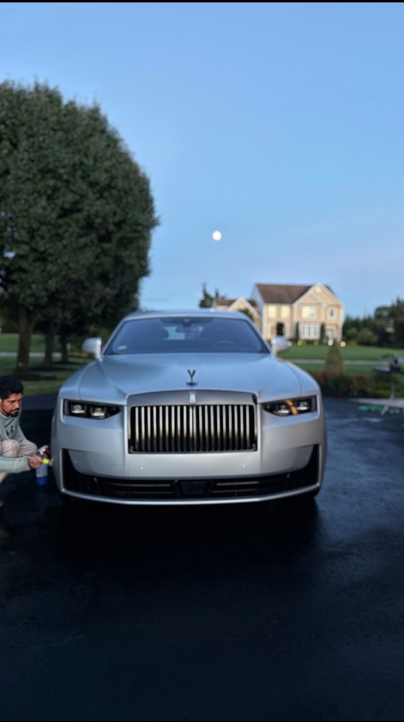 Silver Rolls-Royce parked on a driveway at dusk, with a large tree and house in the background.
