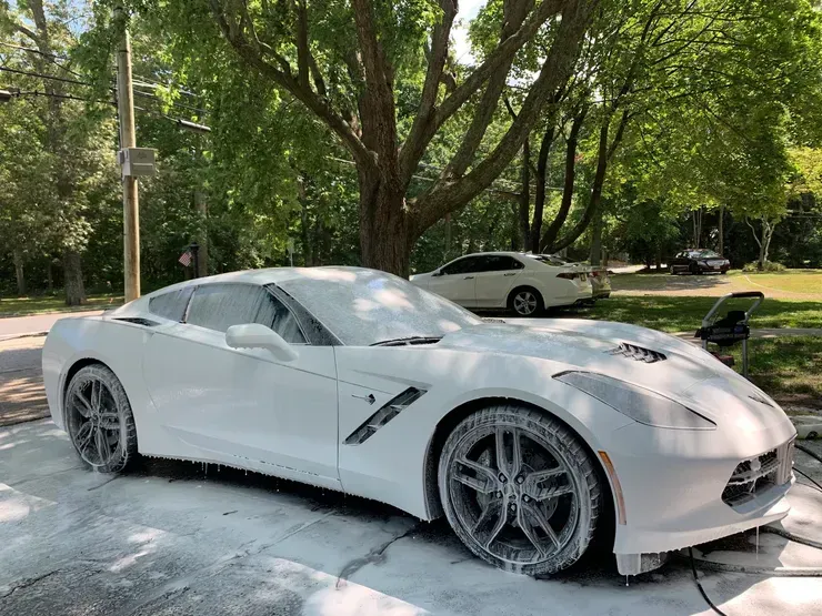 White sports car covered in foam during a wash, parked under trees on a sunny street