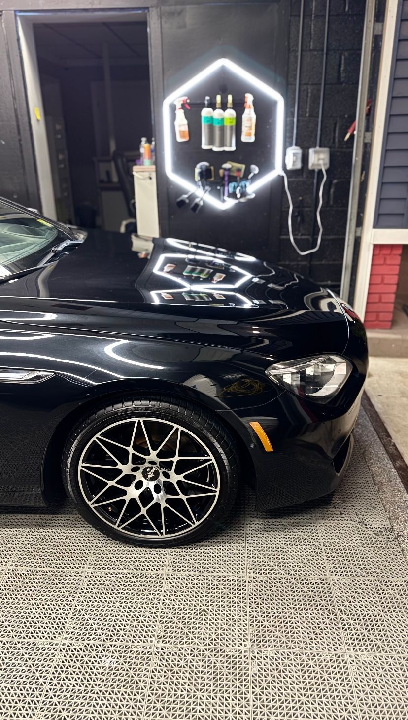 Black sports car parked outside a shop with a neon sign and patterned sidewalk in front.