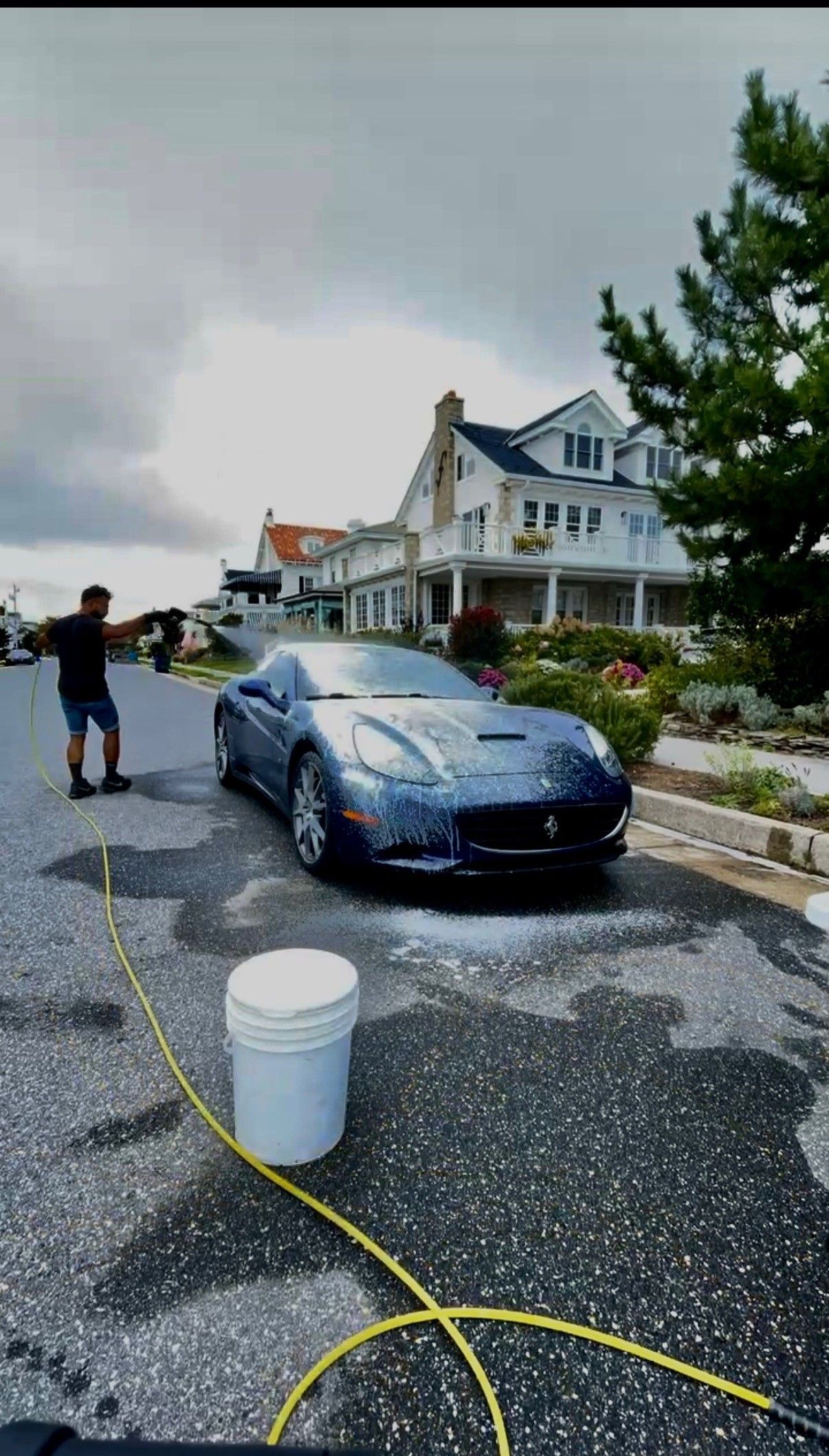 Blue car being washed on a wet driveway outside a large house, with a bucket and hose nearby.