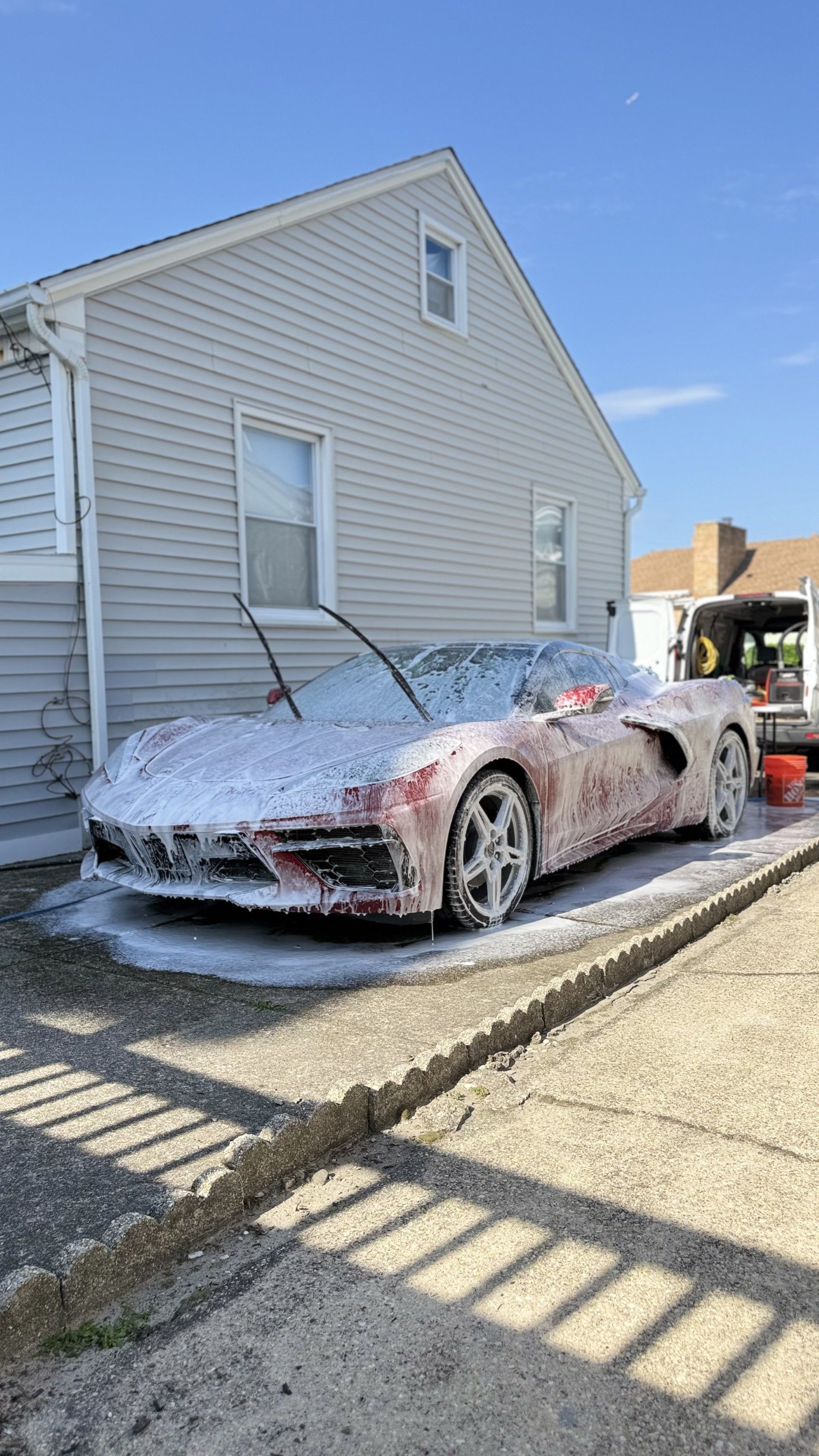 Foam-covered red sports car parked beside a house, partially washed on a sunny driveway.