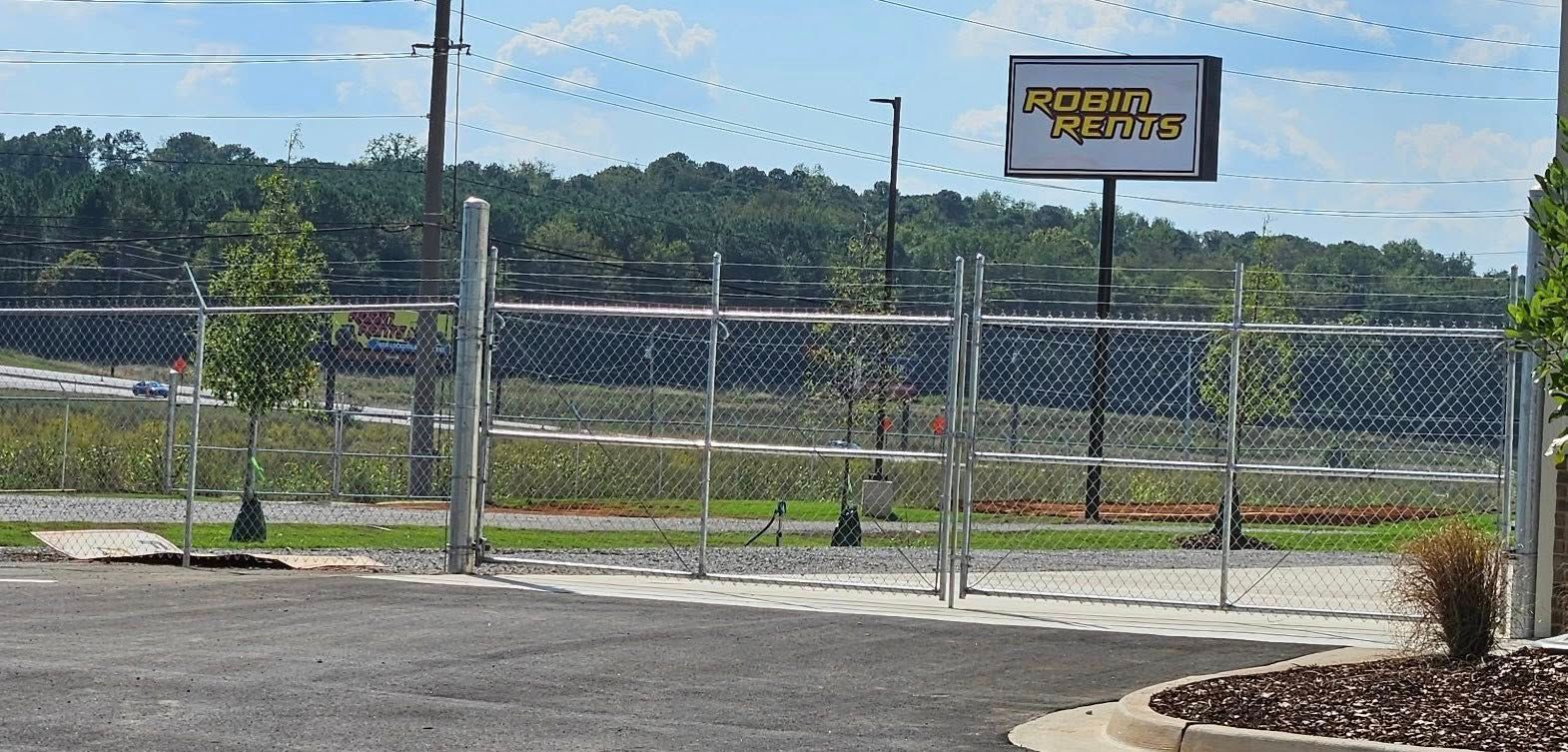 Chain-link fenced lot with a sign, utility pole, and trees under a blue sky