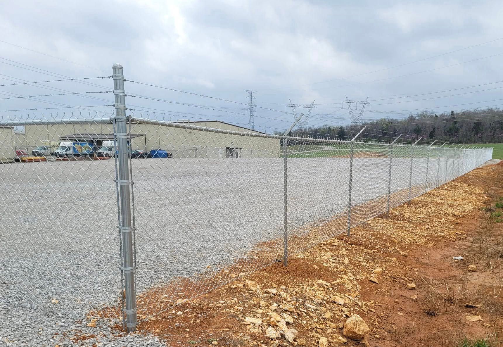 Chain-link fence around gravel lot under cloudy sky, with a dirt road along the right side.