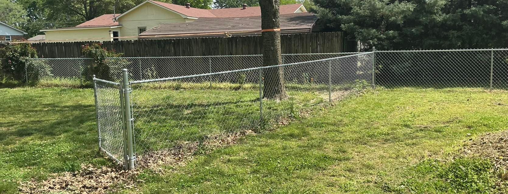 Small fenced garden plot with green grass in front of a house, with a tree in the center.