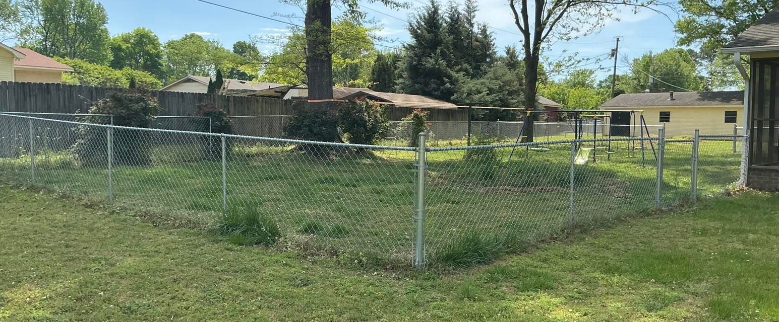 Chain-link fenced grassy lot with trees and houses in the background under a sunny sky