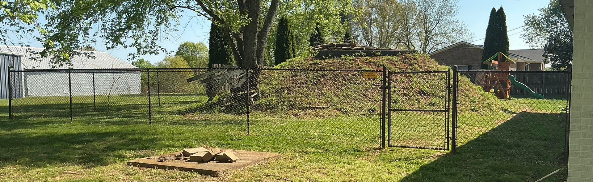 Backyard with a chain-link fence, grassy lawn, and a pile of dirt or mulch under trees.