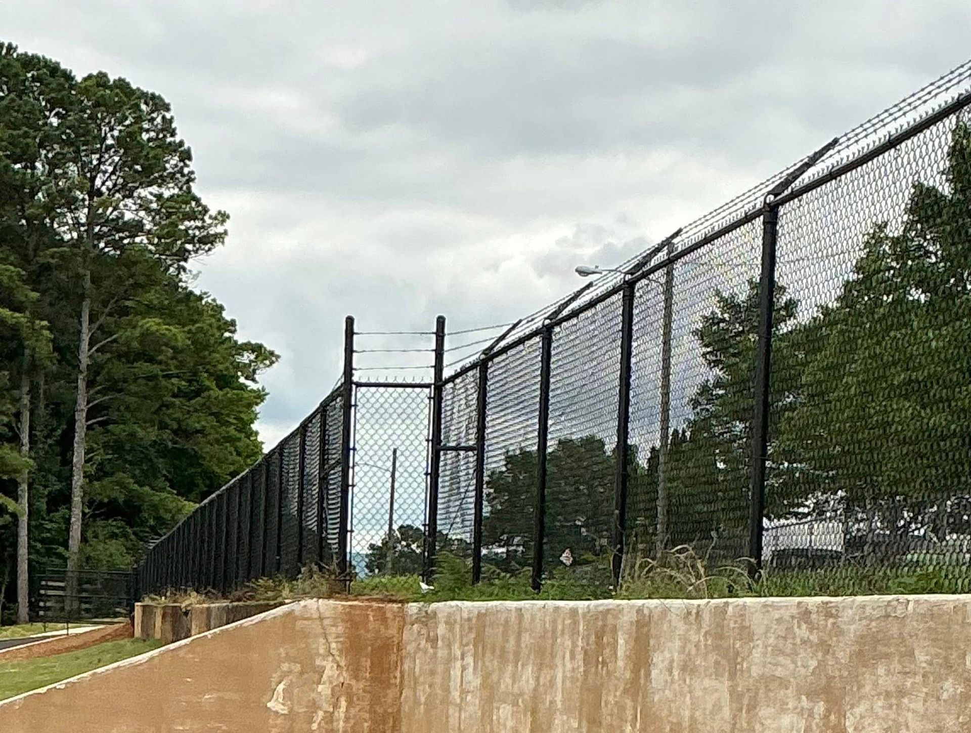 Tall black chain-link fence atop a concrete wall beside a wooded path under a cloudy sky