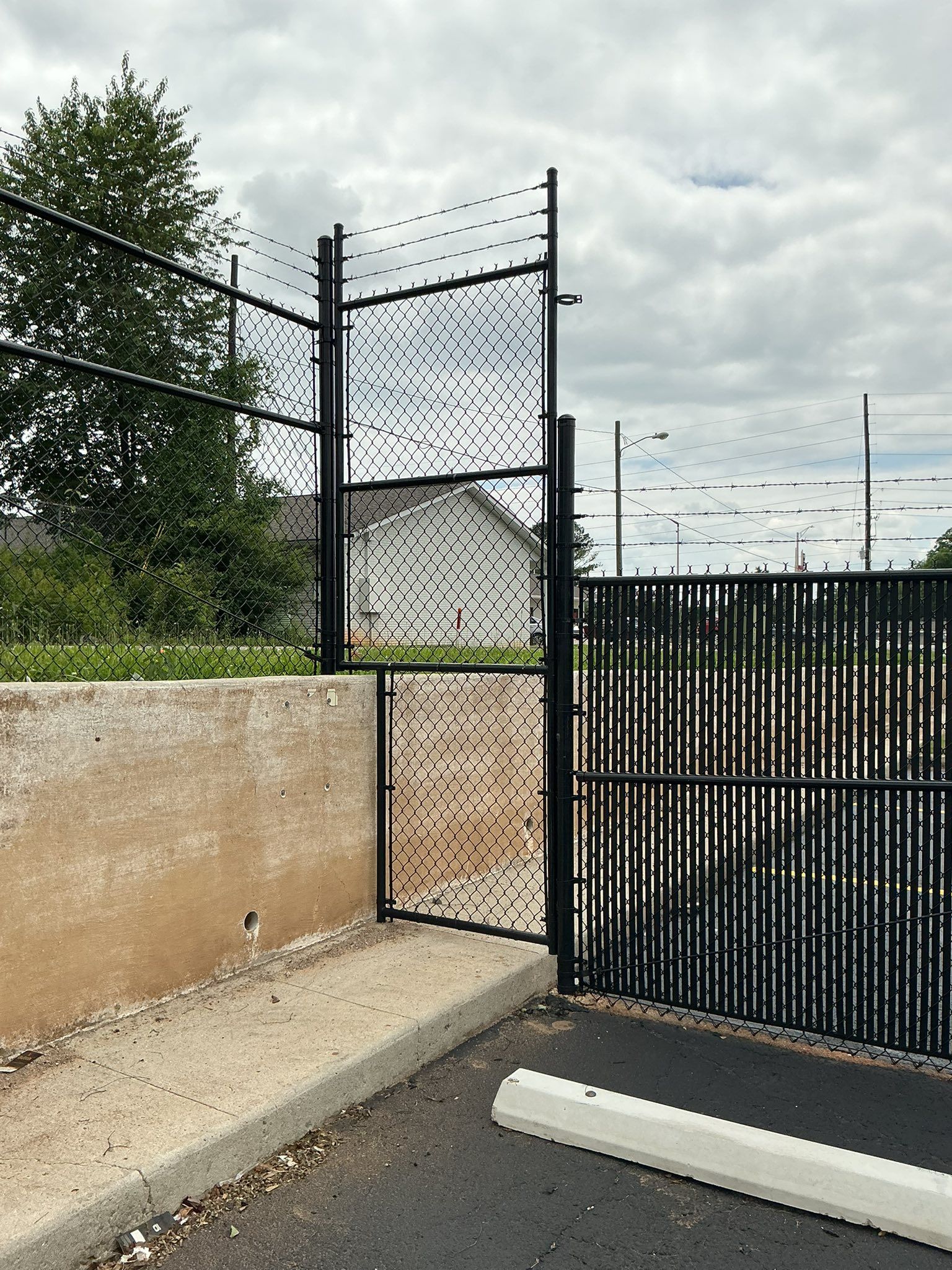Black metal gate beside a beige wall and concrete path, with trees and a house in the background.