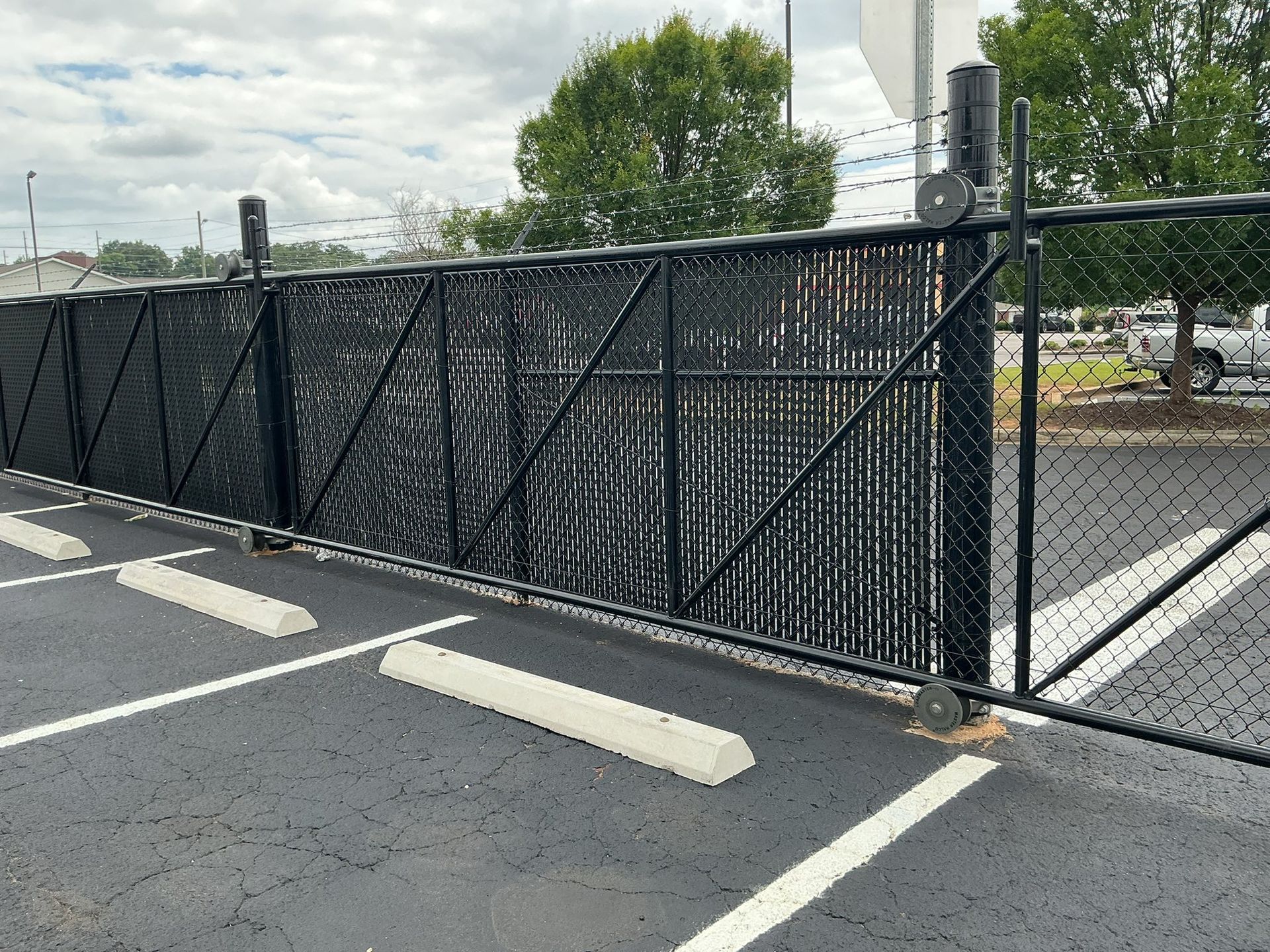 Black chain-link fence and gate blocking a parking lot entrance, with white parking stops and cloudy sky