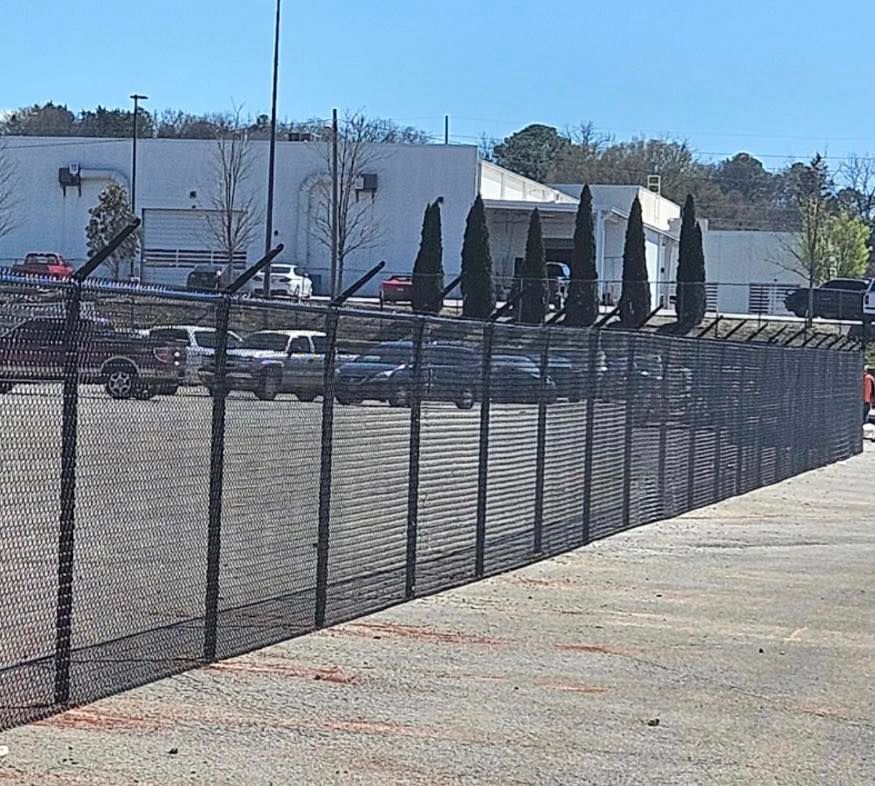 Parking lot with a black chain-link fence, parked cars, and a white building under a clear sky.