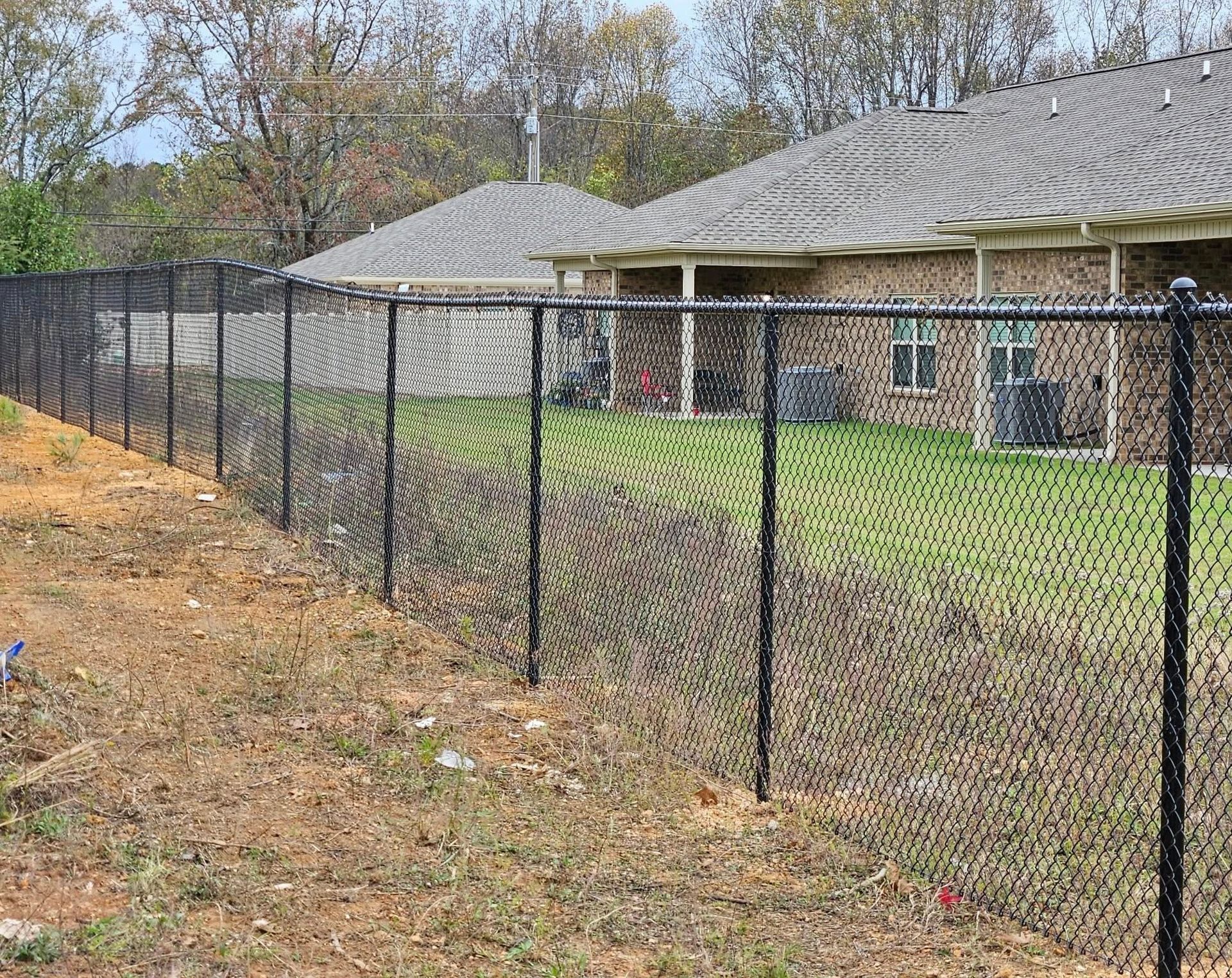 Black chain-link fence beside a brick house with a grassy yard and leaf-covered slope