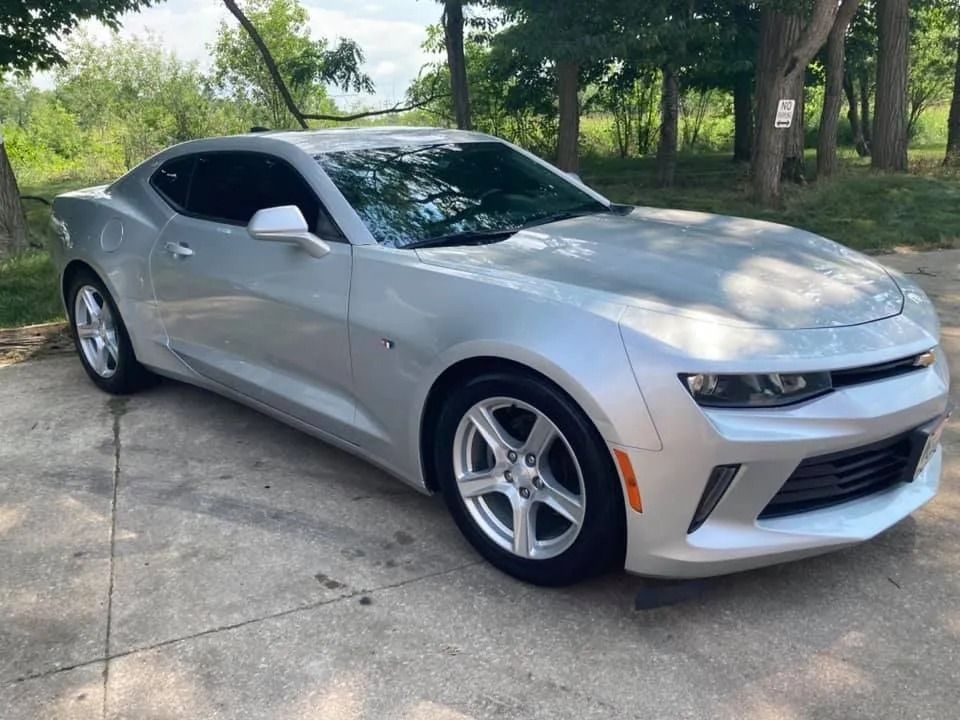 Silver Chevrolet Camaro parked on concrete in front of trees.
