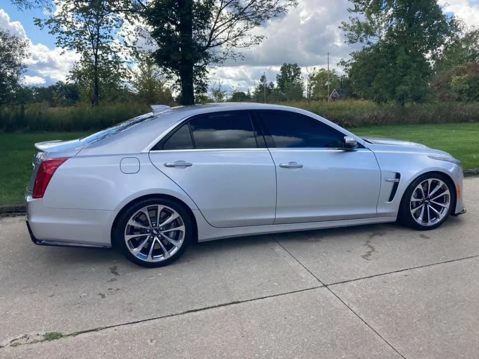 Silver Cadillac CTS-V parked on concrete driveway, side view. Dark tinted windows, sporty wheels, and a rear spoiler.