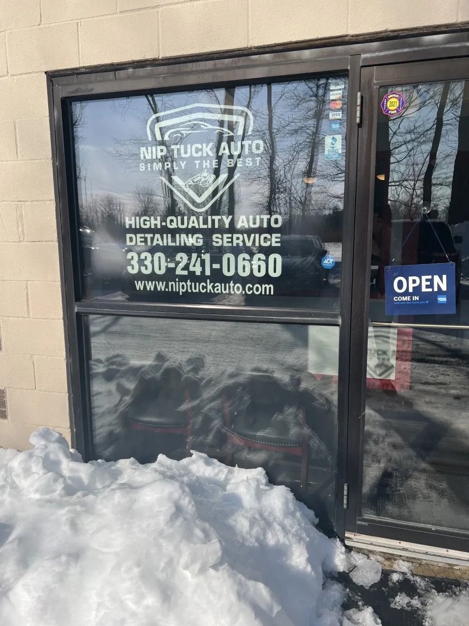 Nip Tuck Auto storefront with business logo, phone number, and website on the window. Snow covers the ground. Open sign visible.