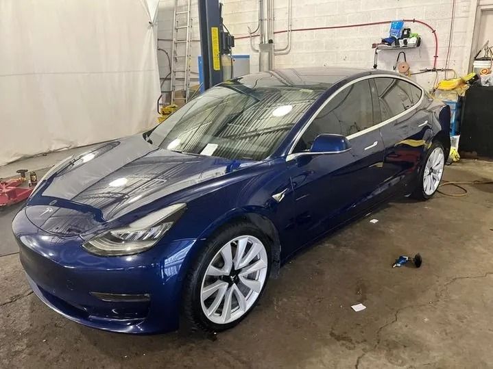 Blue Tesla Model 3 in a garage, shiny paint, white wheels.