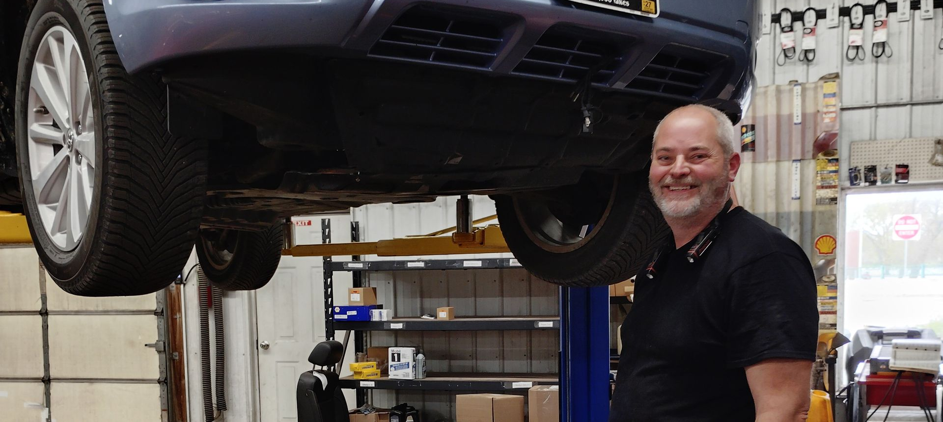 Gray car lifted on a shop hoist, with a smiling mechanic standing underneath in an auto garage.
