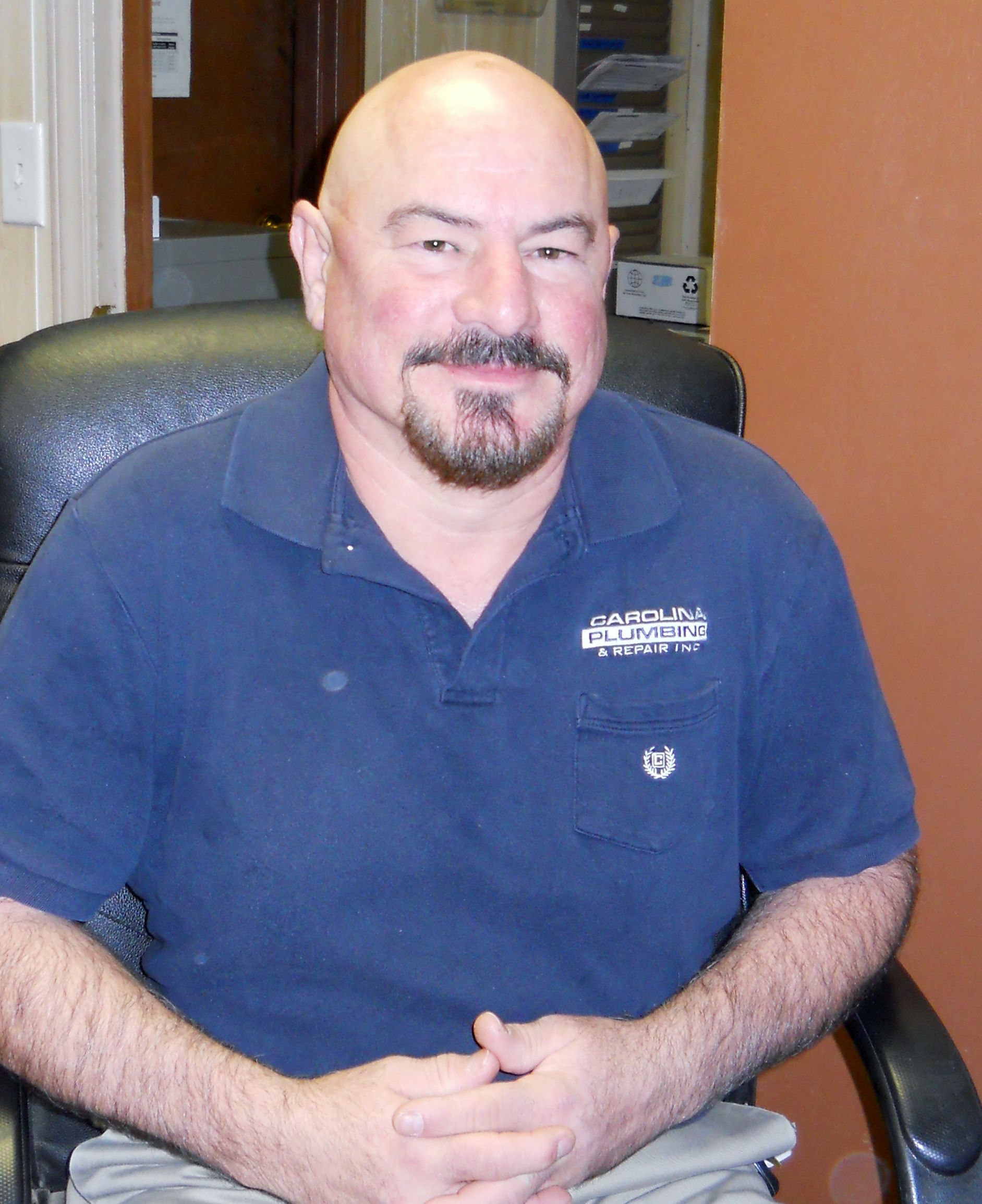 A bald man with a beard is sitting in a chair with his hands folded