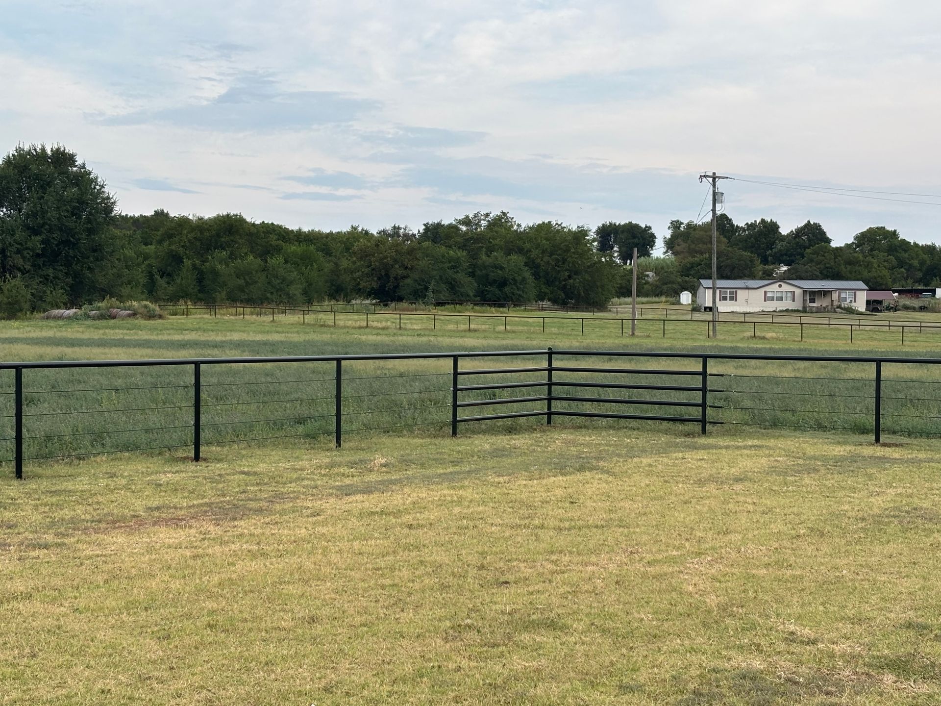 Black fence in a grassy field with trees and a house in the background under a cloudy sky.