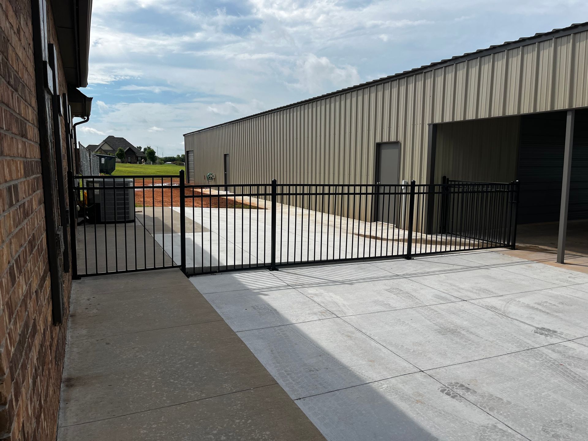 Black metal fence encloses a concrete area next to a brick building and a metal shed under a cloudy sky.