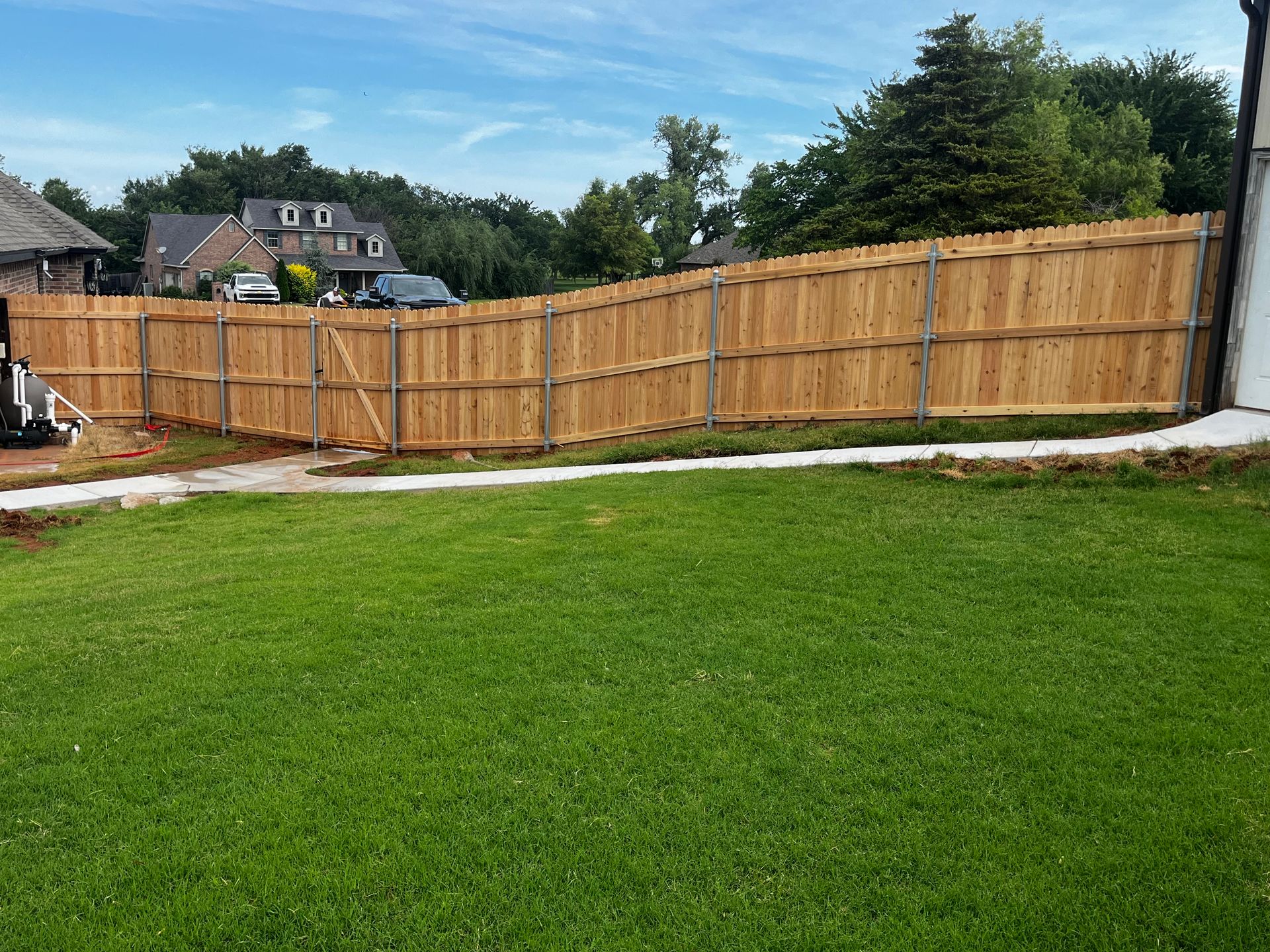 A wooden fence curves around a grassy yard under a blue sky. Houses and trees are in the background.