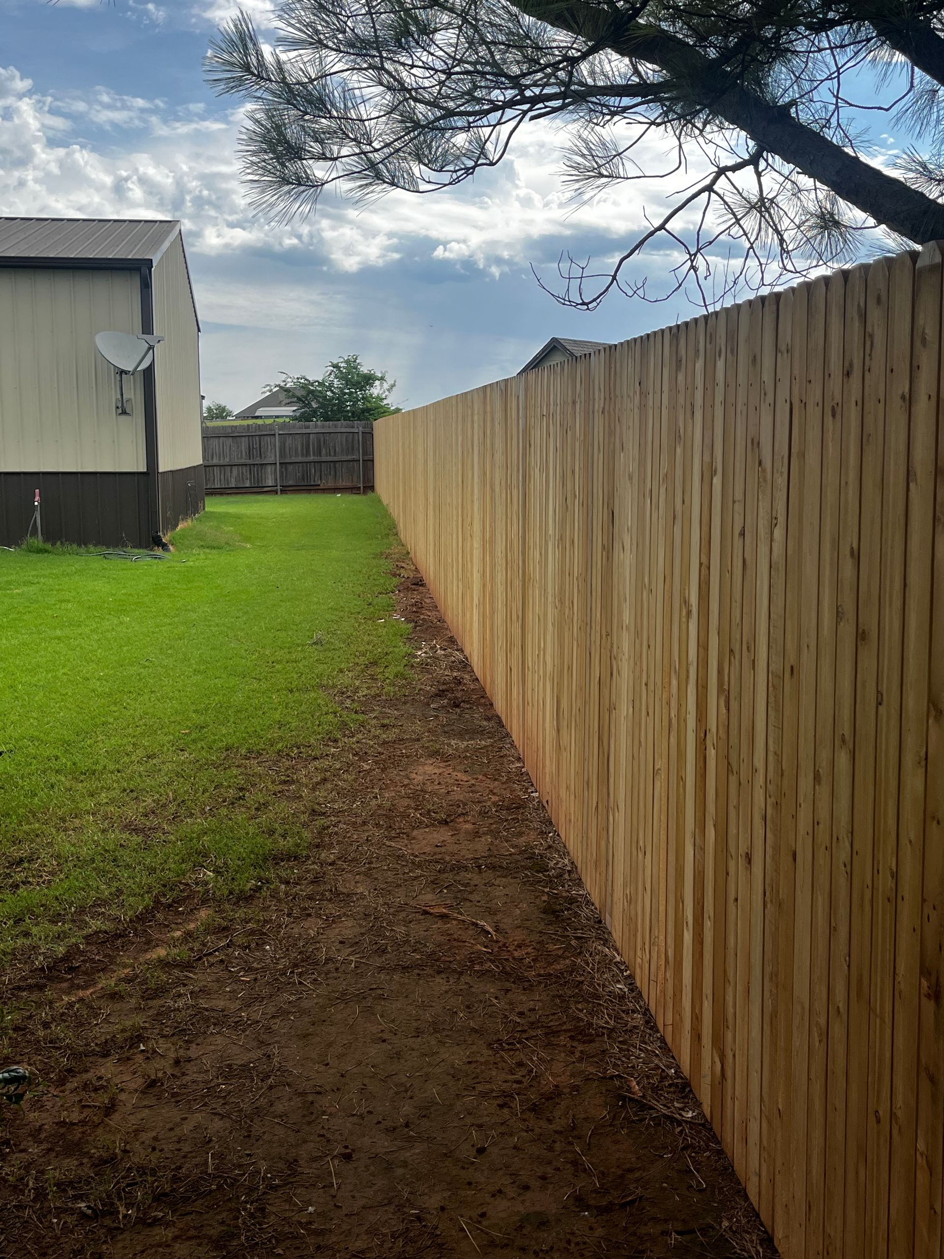 A freshly-built wooden fence runs alongside a green lawn and a house on a cloudy day.
