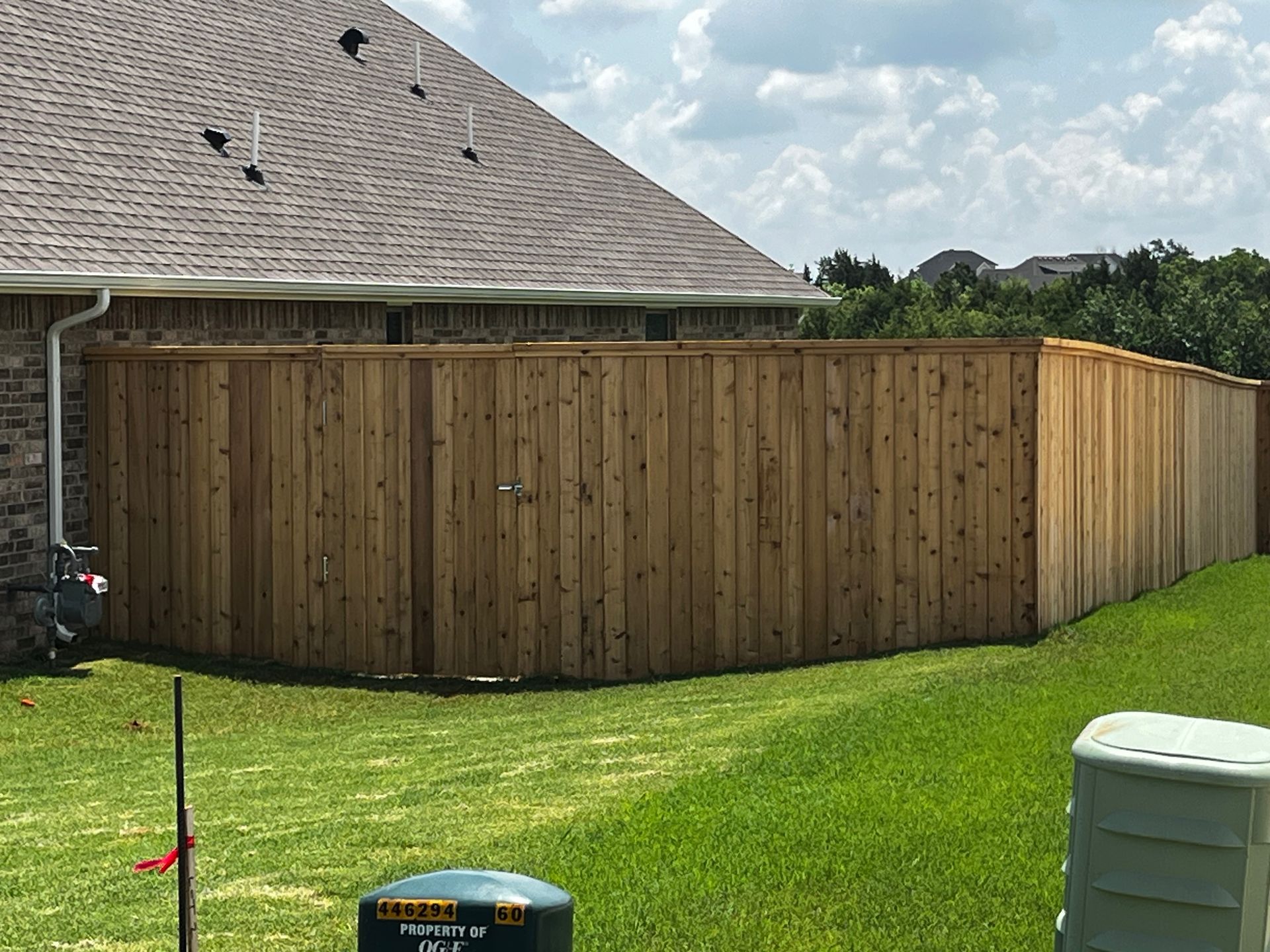 A wooden fence surrounds a grassy backyard next to a brick house under a cloudy sky.