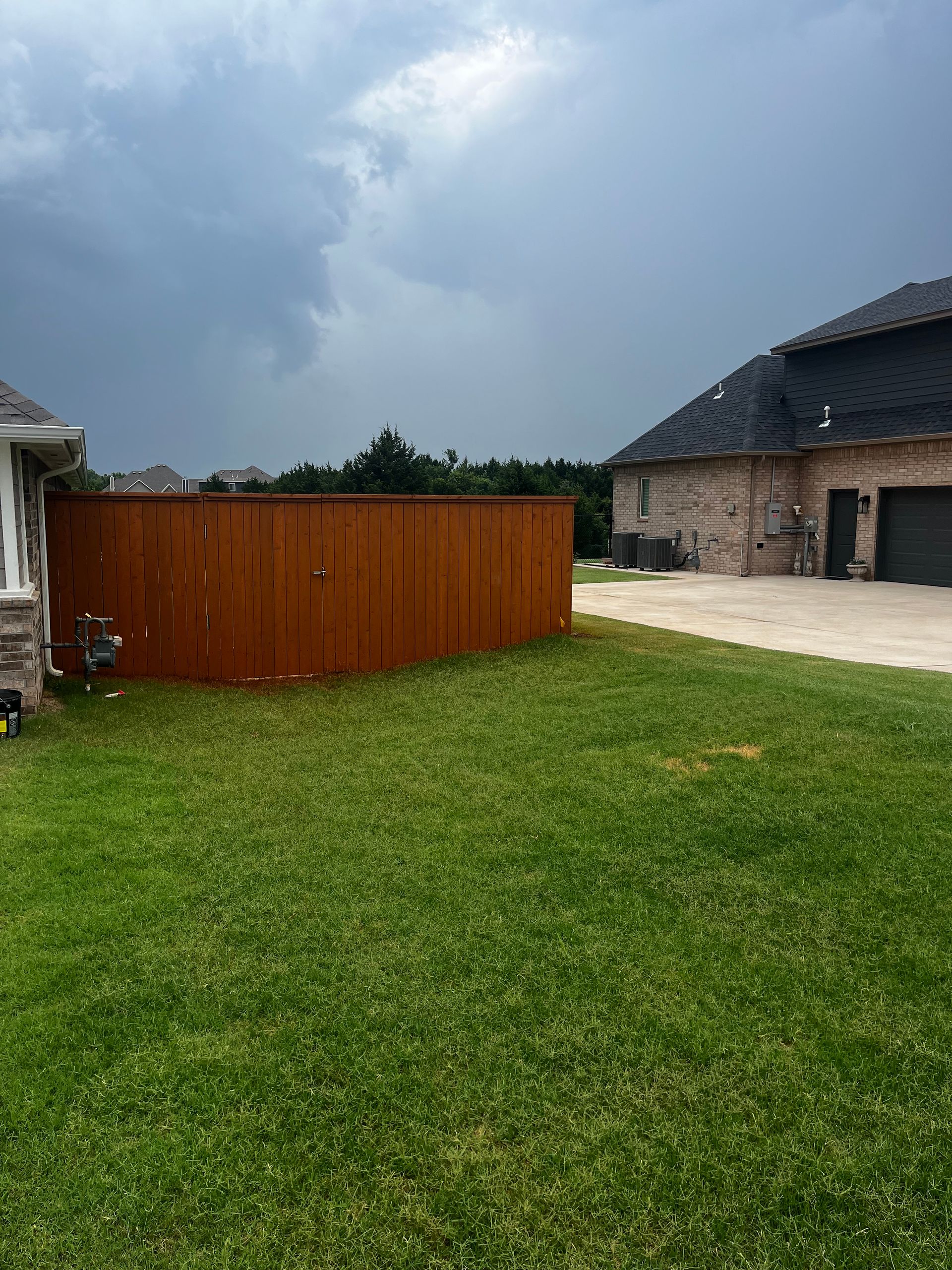 A wooden fence in a yard under a cloudy sky. Grass in the foreground, brick house in background.