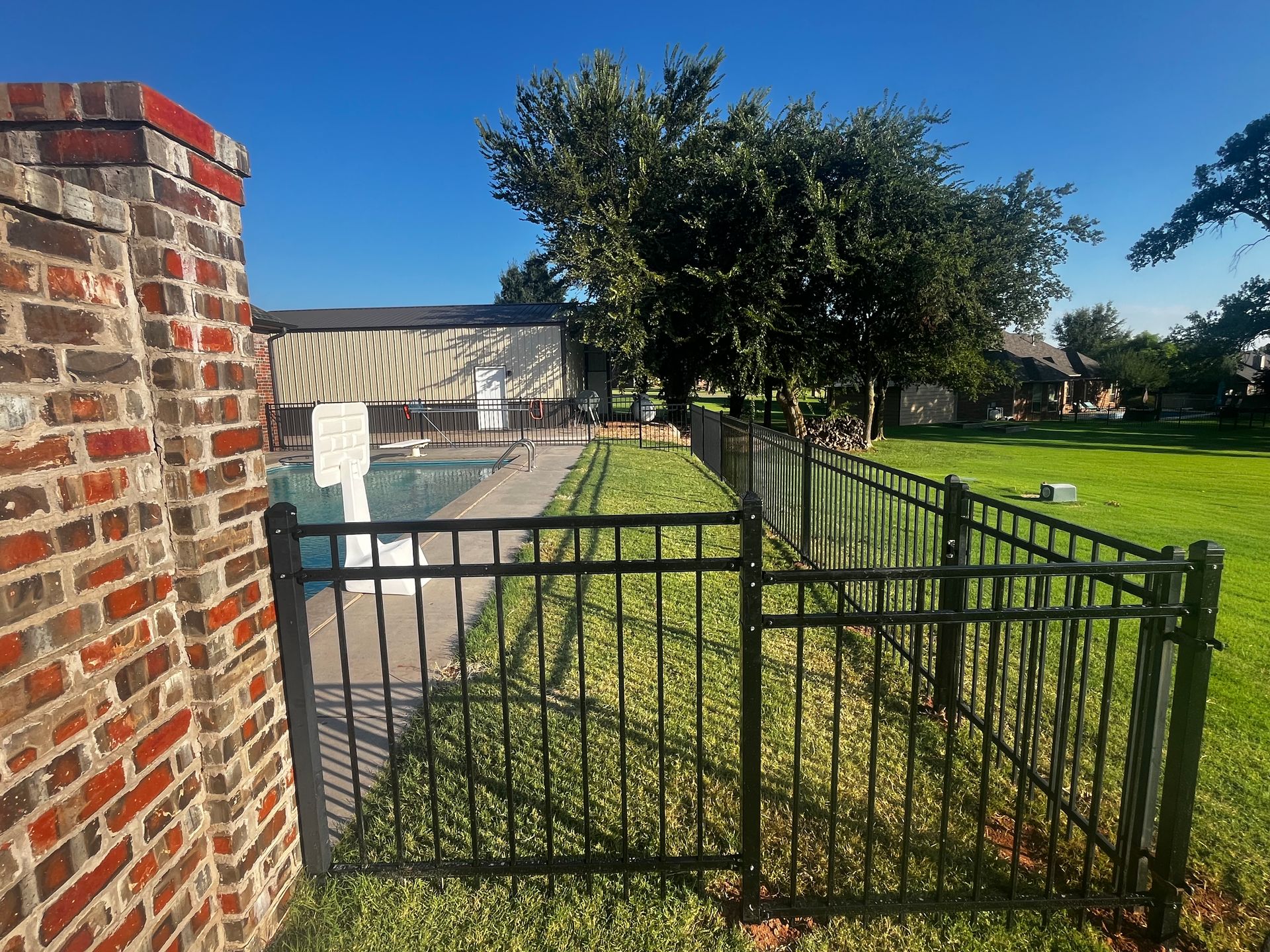 Black fence surrounds a pool and lawn, brick wall on the left, trees and building in the background, clear blue sky.