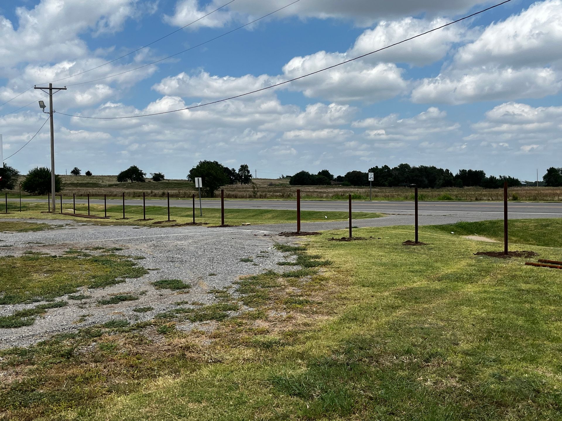 Open field with a gravel parking area and metal fence posts under a cloudy sky.