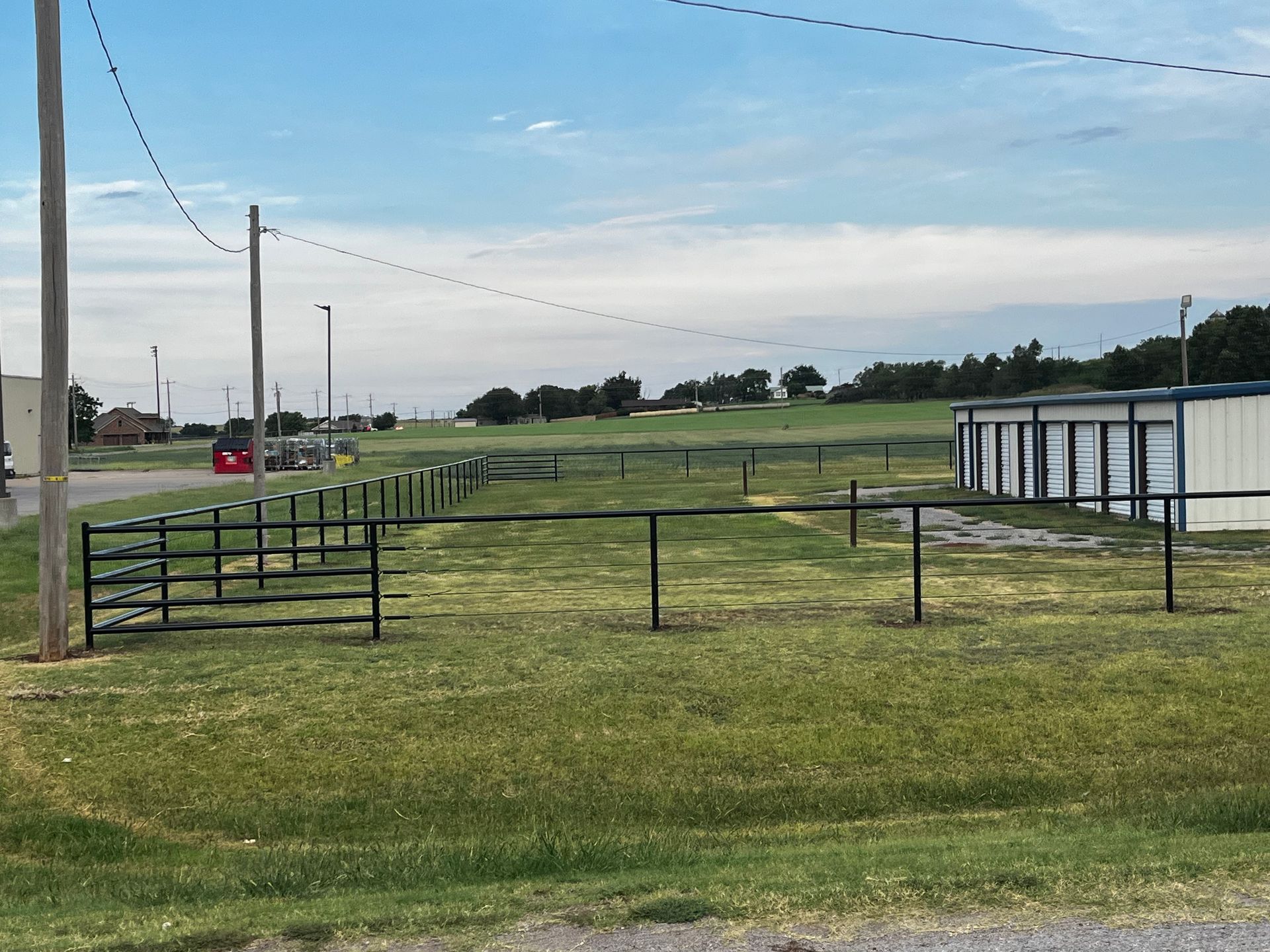 Grassy field with black metal fences, power lines, and storage units under a blue sky.