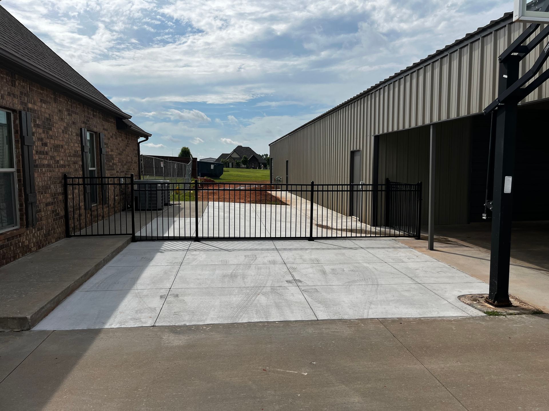 Black metal gate on a concrete driveway between a brick house and a metal building.