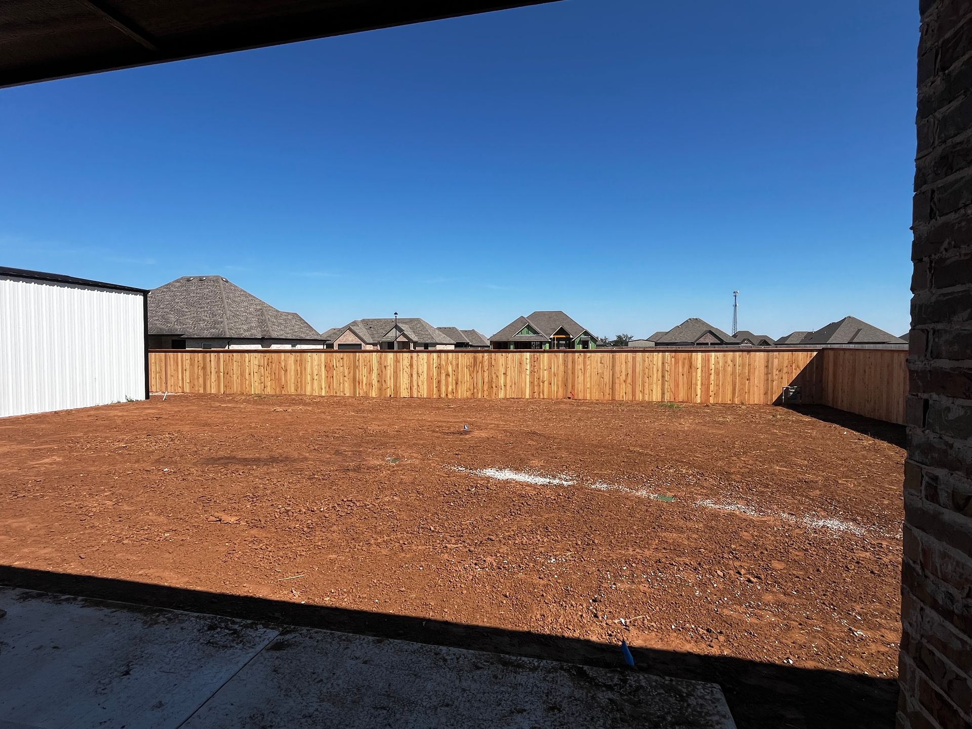Backyard view of wooden fence, clear blue sky, houses in the distance.