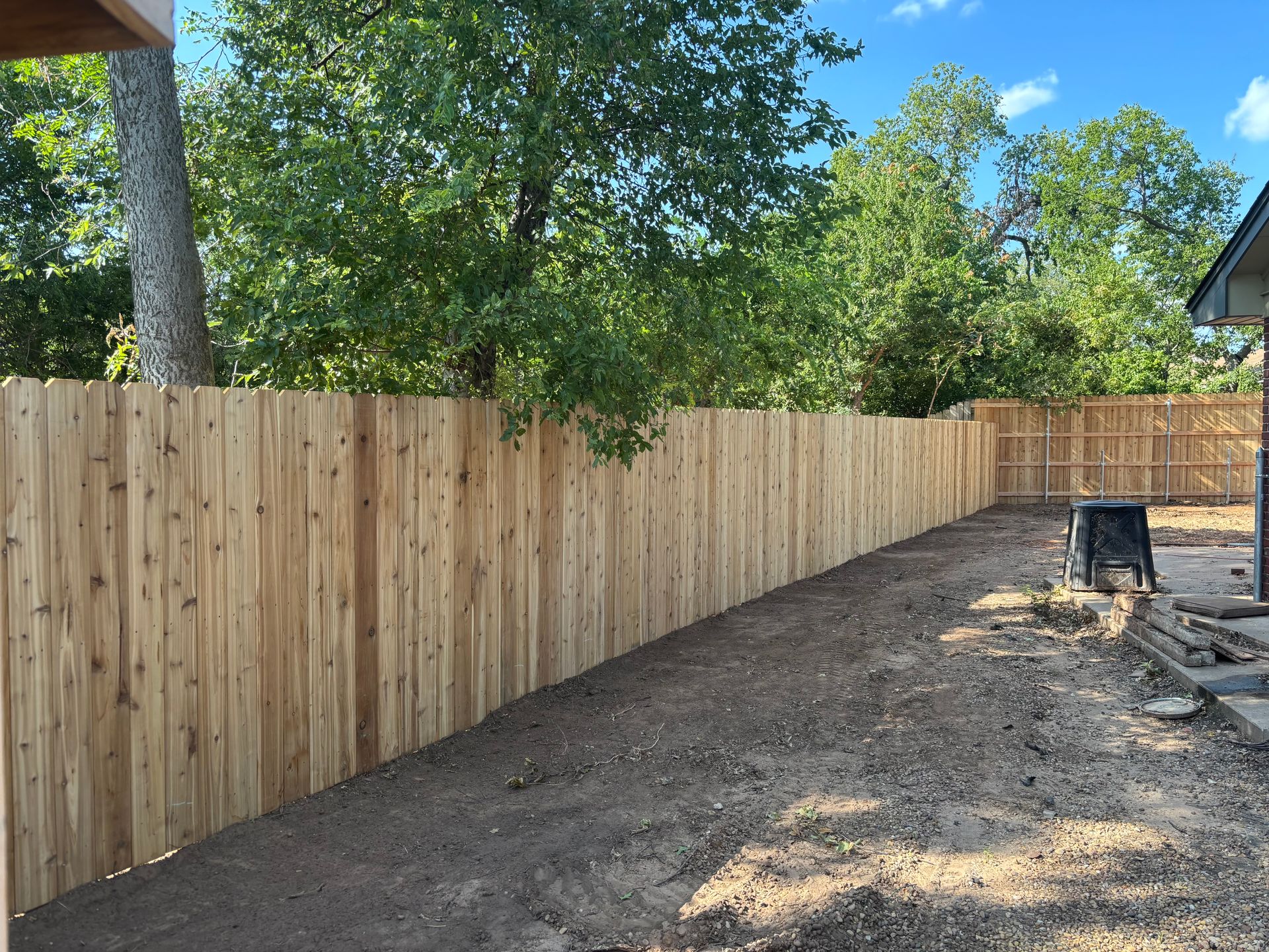 Wooden fence in a backyard, with dirt ground and trees in the background under a blue sky.