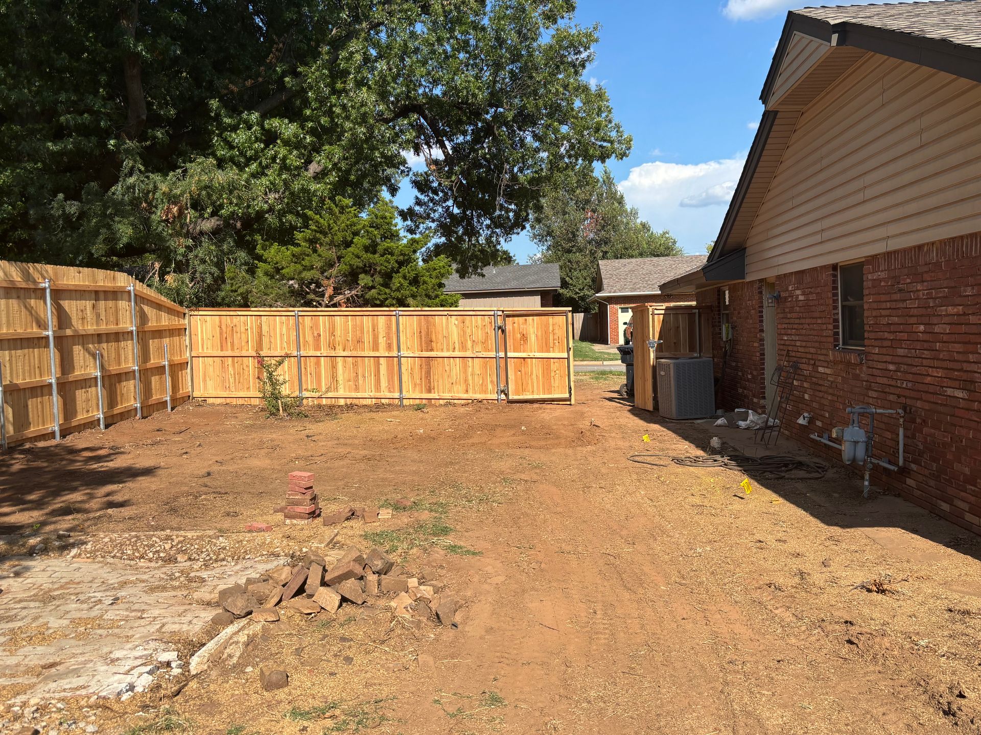 A newly fenced backyard with dirt ground, a brick house, and wooden fence under a blue sky.