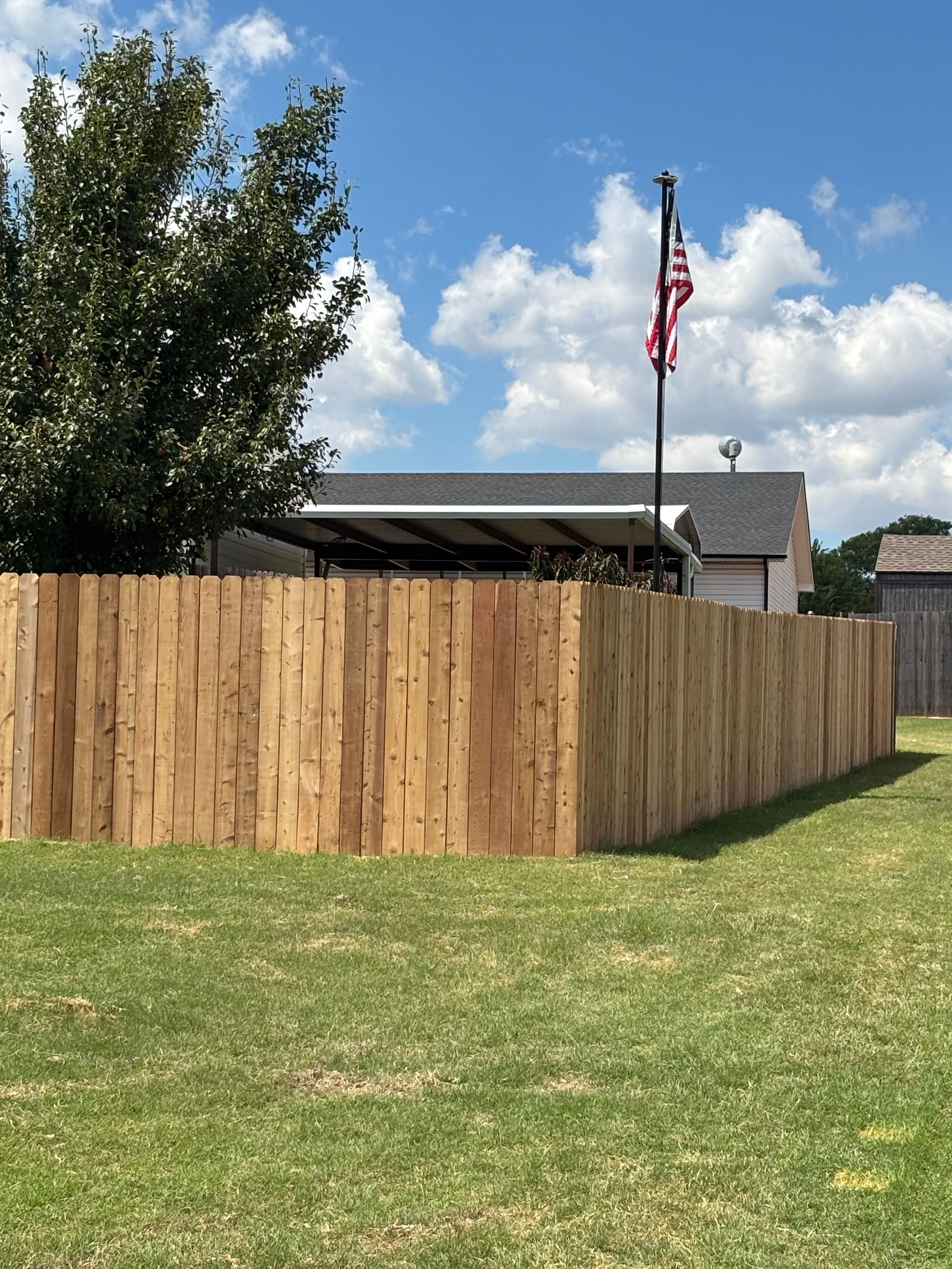 Wooden fence in a grassy area, with a building and American flag in the background under a blue sky.