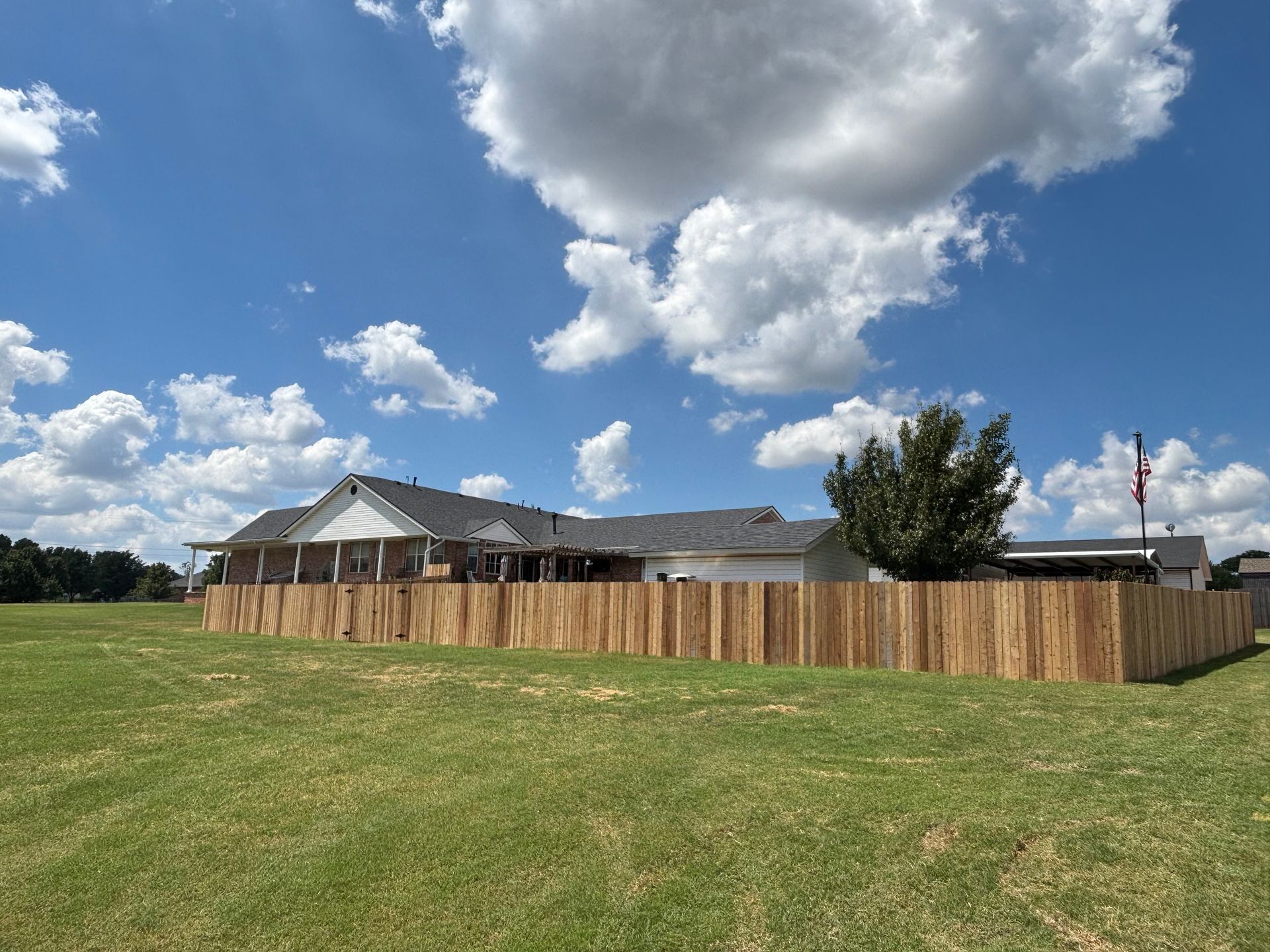 A wooden fence surrounds a house on a grassy field under a blue sky with fluffy clouds.