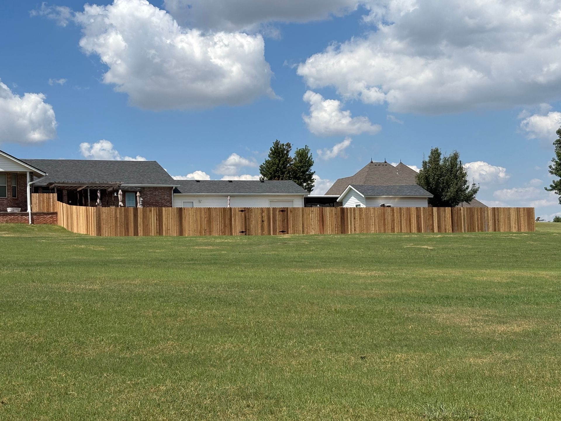 Wooden fence in a grassy yard, with houses and a blue sky with clouds in the background.