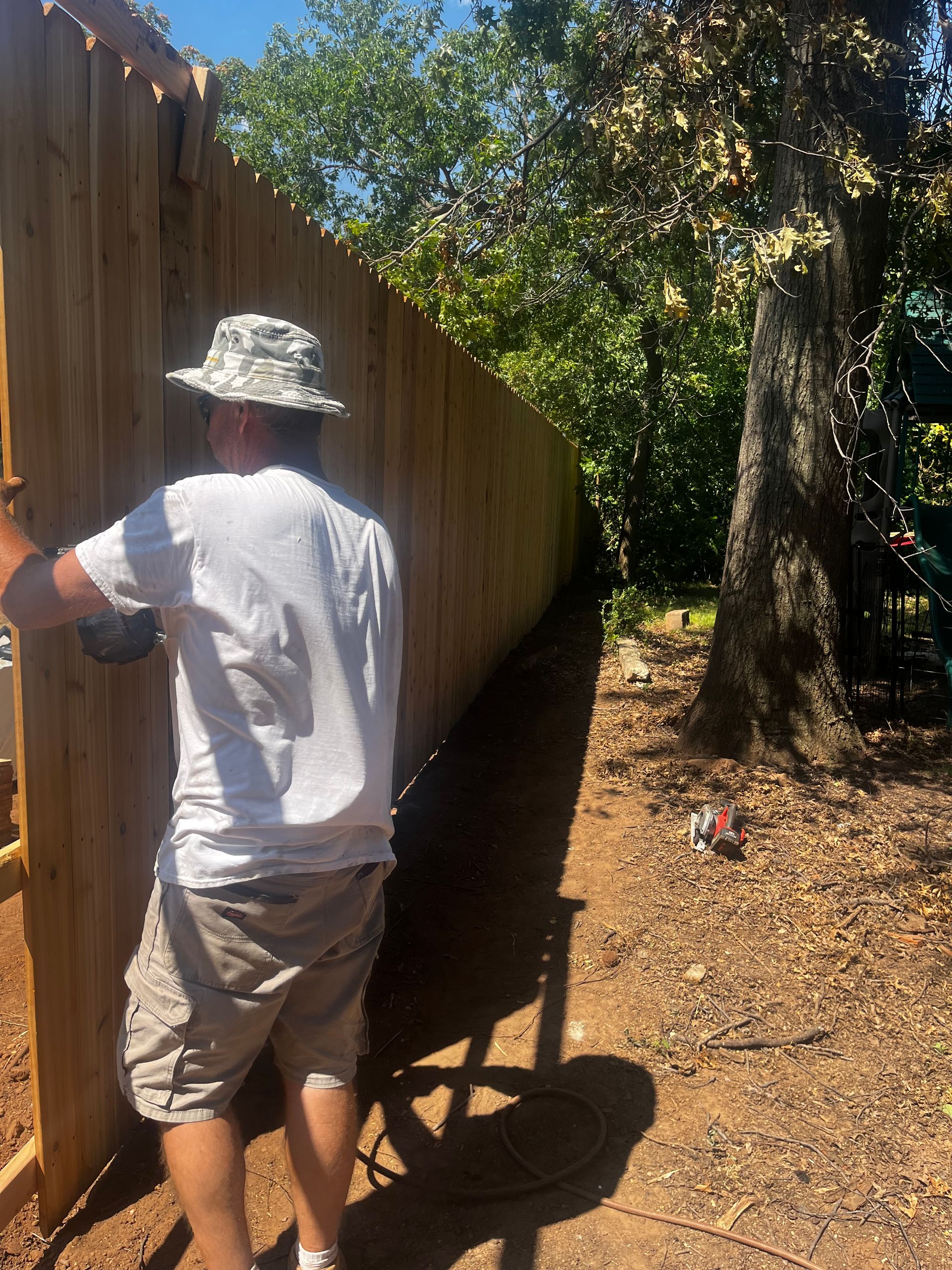 Man in hat building wooden fence in sunny yard.