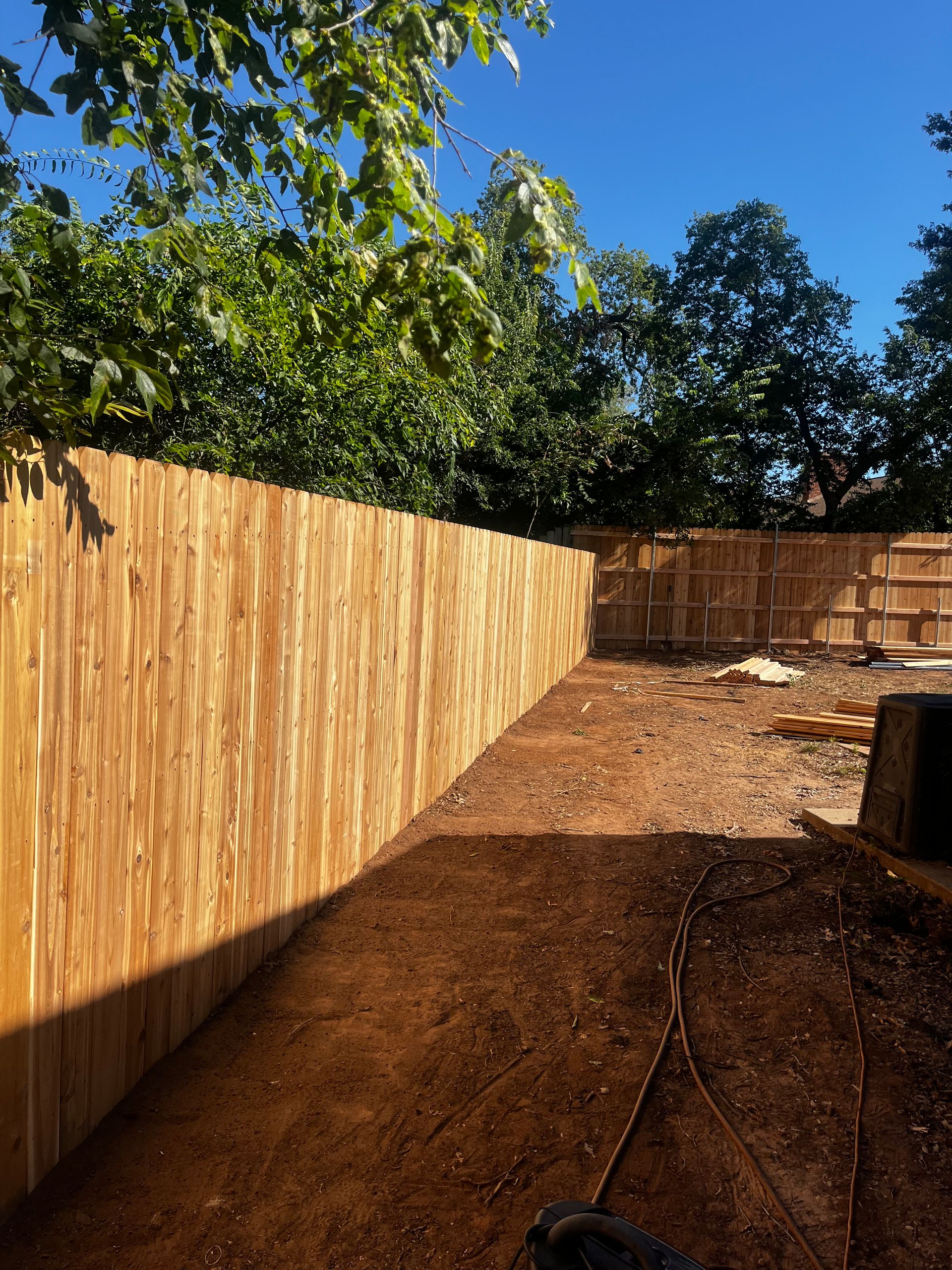 Newly built wooden fence enclosing a dirt backyard, trees in the background, sunny day.