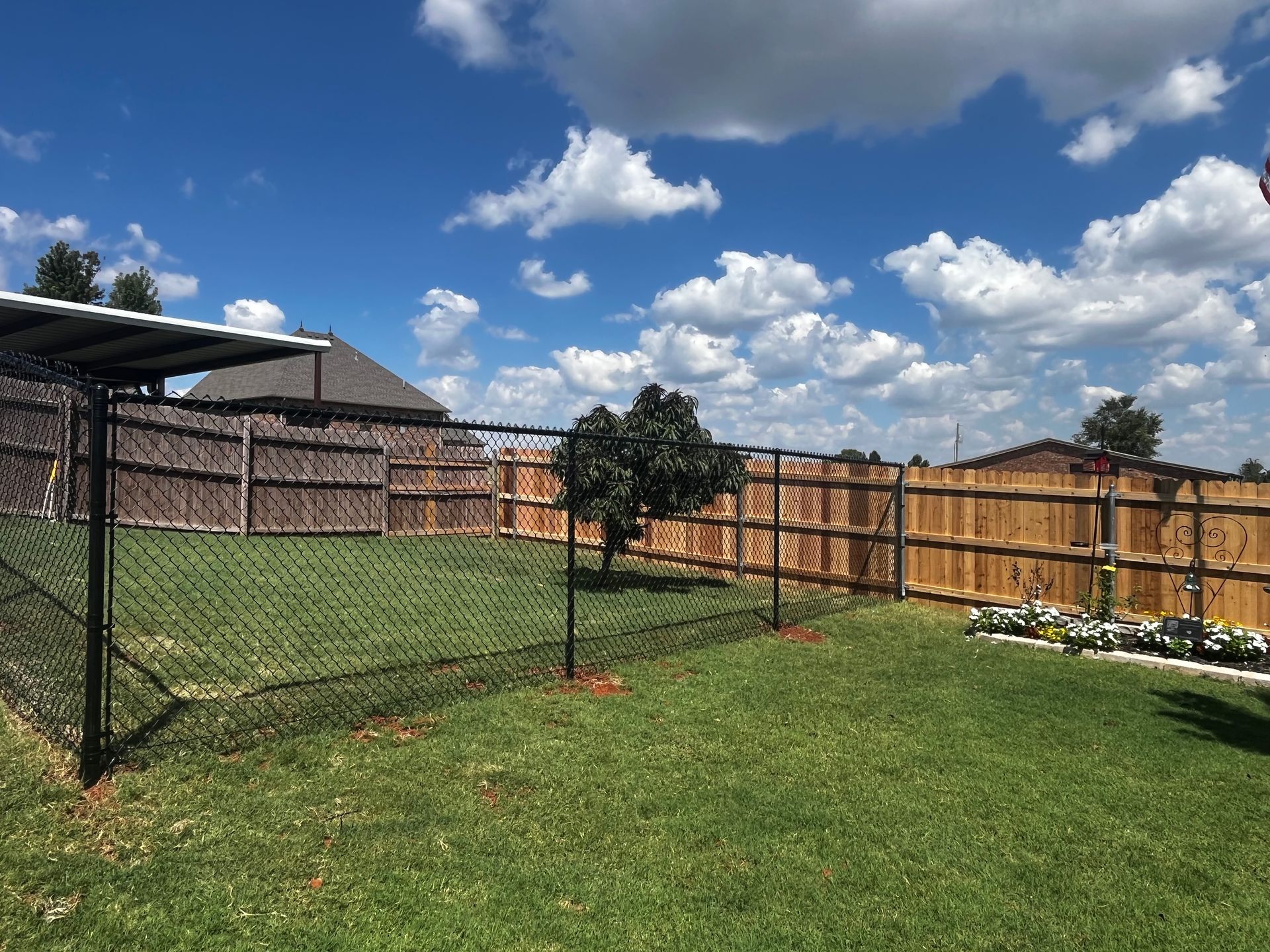 Lawn with chain-link fence, wooden fence, blue sky with clouds, small tree, and shade structure.