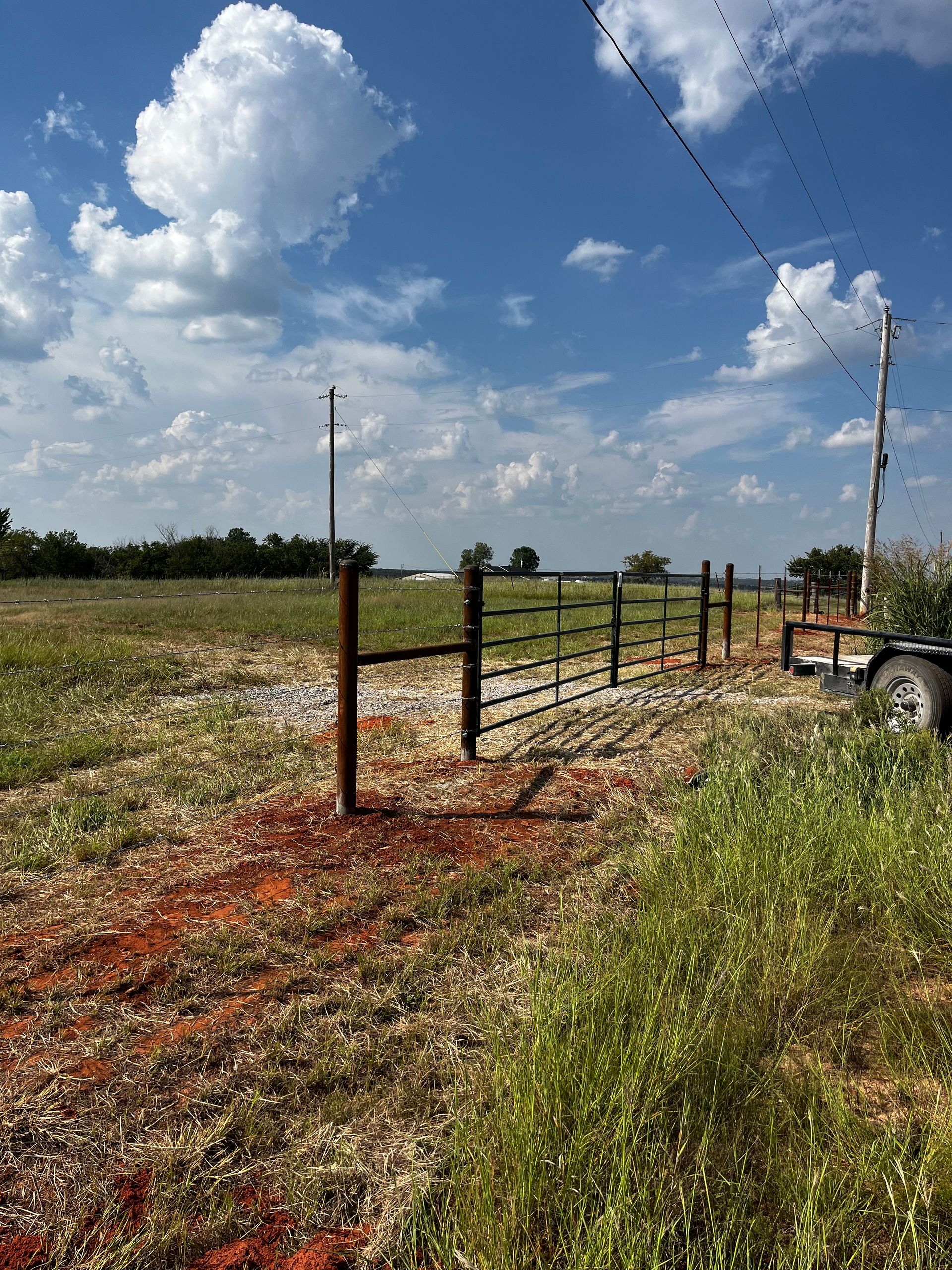 A metal gate in a grassy field under a bright blue sky with fluffy clouds, telephone poles, and a trailer.