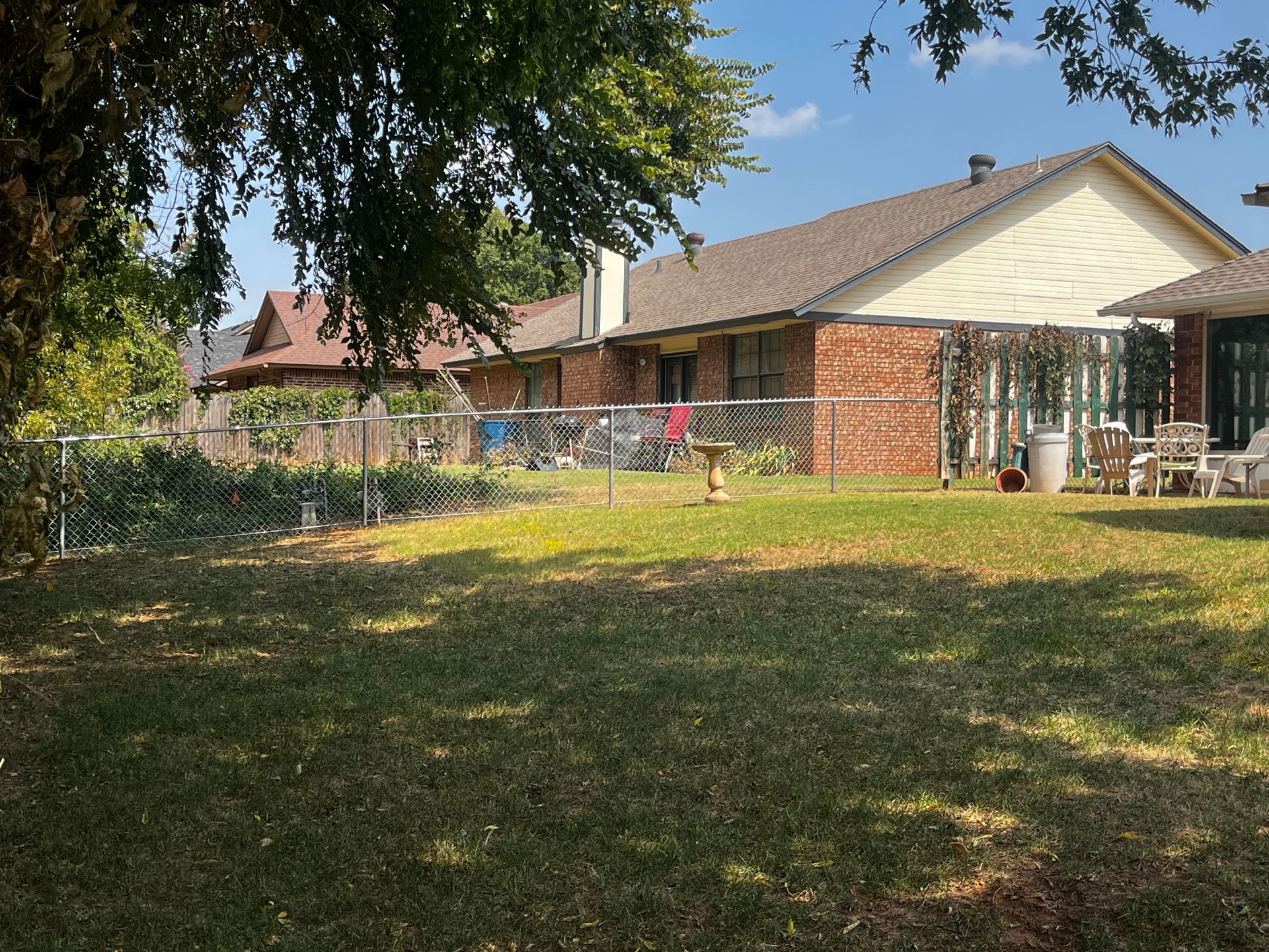 Backyard with grass, a chain-link fence, and a brick house under a sunny sky.