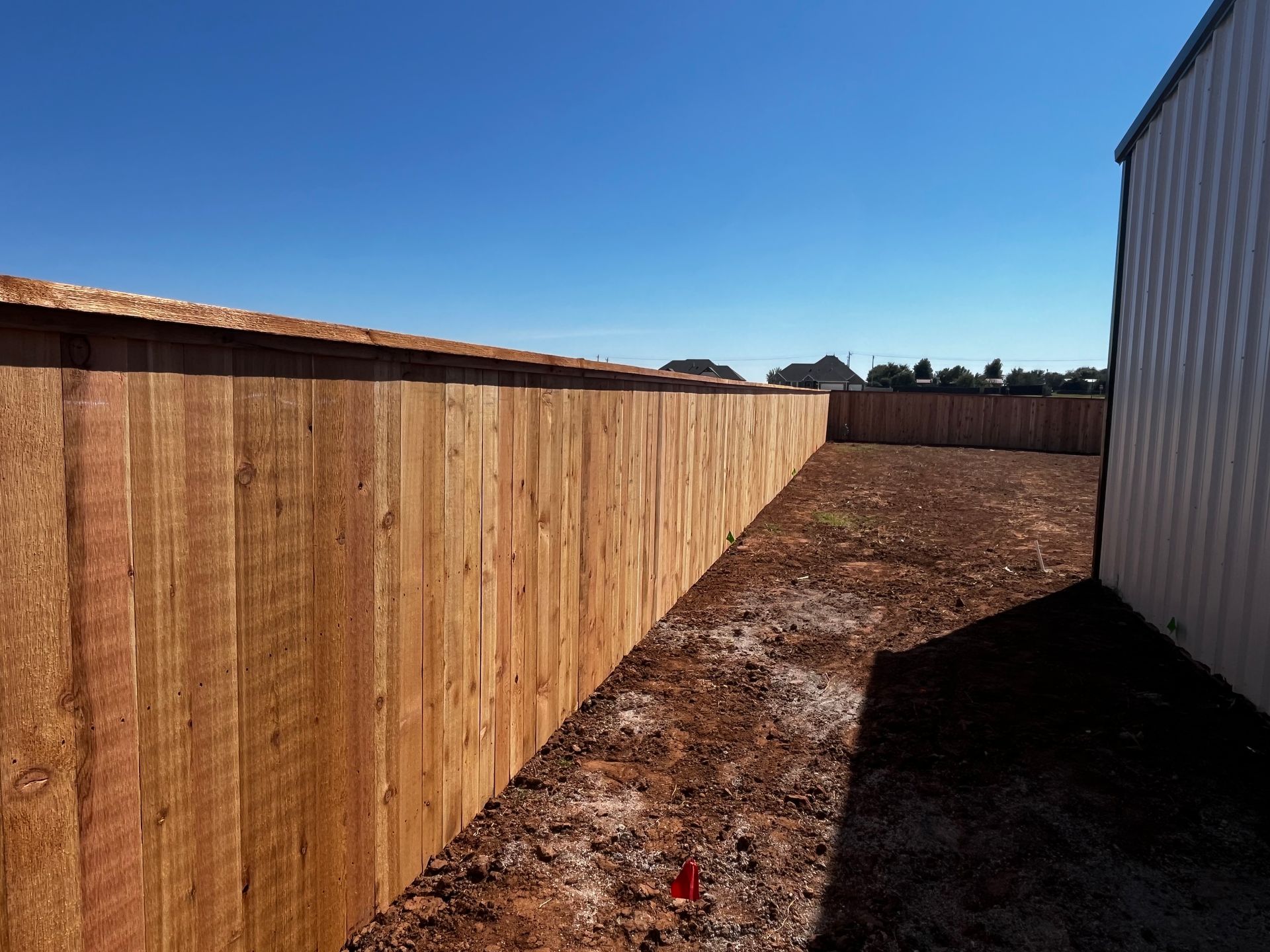 Wooden fence in an empty dirt lot on a sunny day, with a building visible on the right.